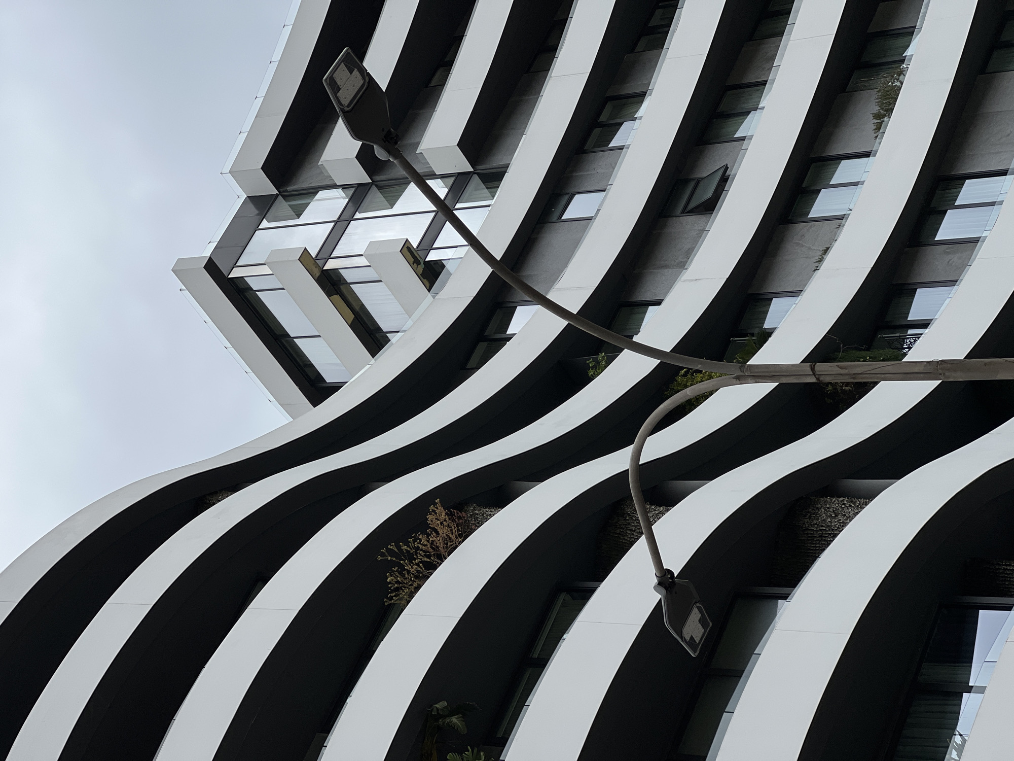 Here is a detailed description of the image:
The image shows a low-angle view of a modern building with a striking architectural design, consisting of alternating black and white horizontal bands that create a wave-like pattern. Windows are visible within the black bands. Plants and greenery are nestled in the structure, suggesting an effort to integrate nature into the building's design.
A street lamp is prominently featured, with two lights extending from a curved pole.
The sky is overcast, contributing to a somewhat muted color palette.
Based on the architectural style and the presence of distinctive design elements, it is possible that this building is located in a contemporary urban setting, possibly Budva, Montenegro.