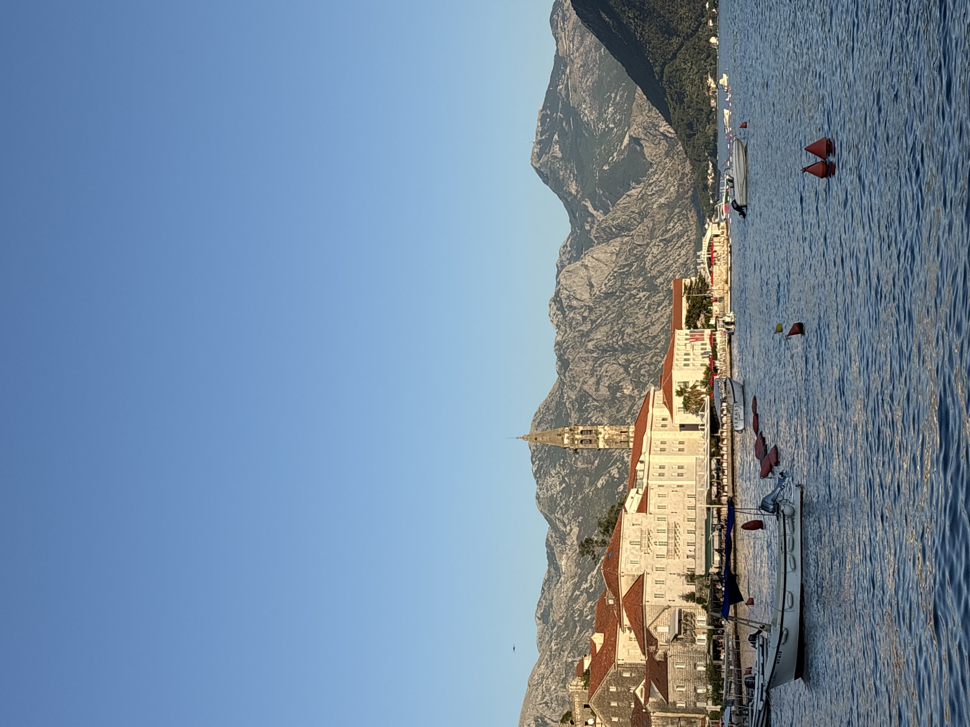The image depicts a serene waterfront scene in Perast, Montenegro, under a clear blue sky. In the foreground, calm blue water reflects the sunlight, with several small boats anchored or moored. Some boats are white, and one has an outboard motor. Red buoys are visible floating on the water, marking an area or pathway.
Along the shore, a row of stone buildings with distinctive red-tiled roofs forms the town's facade. The architecture appears historic, with arched windows and classical detailing. A prominent bell tower, a common feature of European coastal towns, rises above the buildings, its stonework weathered by time. Beyond the town, a range of imposing, rugged mountains forms a dramatic backdrop, their rocky slopes covered in sparse vegetation. The overall impression is one of a picturesque and peaceful European coastal town.
