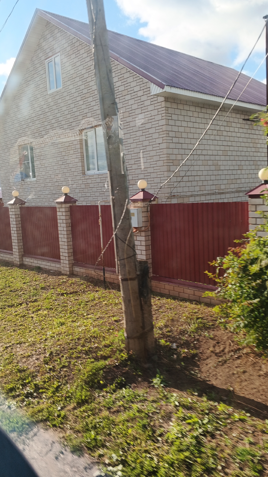 A side view shows a light-colored brick house with a dark red metal roof and matching metal fence. The house has two visible windows on the upper level and one on the lower level, all with white frames. The fence is made of corrugated metal panels and is supported by brick pillars topped with decorative caps and lights. A utility pole stands in front of the fence, with wires running from it. The ground in front of the fence is covered in grass and dirt. The scene appears to be in a suburban or rural area, possibly in the city of Kiasovo, Russia, given the context. The overall impression is one of a well-maintained property.