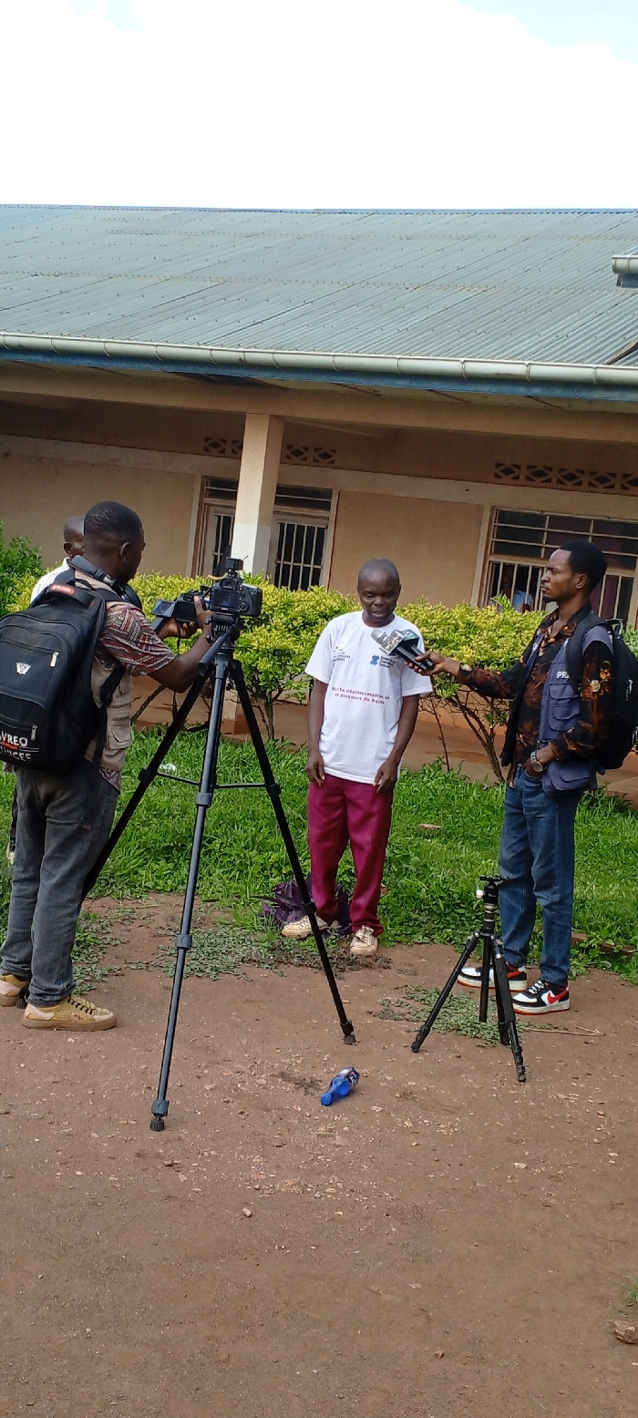 The scene depicts an outdoor media recording session involving four individuals in Kundu 1, Democratic Republic of the Congo. The ground consists of reddish-brown dirt and gravel with scattered green vegetation.
From left, a male cameraman, wearing a patterned shirt and dark trousers, operates a black professional video camera mounted on a tall black tripod. A dark backpack with "AVREO SCIENCES" text is visible on his back. Partially obscured behind him is another male figure in a light-colored shirt. Centrally, a male interviewee, appearing older, wears a white short-sleeve t-shirt with blue and black logos, featuring the French text "Non à la désinformation et au discours de haine." He wears dark red trousers and light-colored sneakers. To the right, a male interviewer, dressed in a multi-patterned shirt, blue jeans, and white, black, and red sneakers, holds a microphone with a blue foam windscreen and "RTNC" branding towards the interviewee. He carries a dark backpack labeled "PRESSE." A smaller dark camera on a compact black tripod is positioned near the interviewer's feet. A blue plastic bottle lies on the ground between the two tripods.
The background features a light-brown colored building with visible windows secured by bars and supporting columns. Green shrubs are planted in front of the building. The structure is topped by a gray corrugated metal roof equipped with a light blue gutter. The sky above is light blue with sparse white clouds.