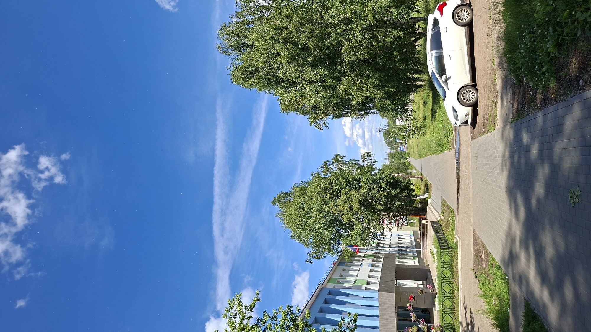 The image depicts a wide pedestrian walkway in a residential area of Chaikovsky, Russia. The walkway is paved with grey interlocking concrete pavers and runs parallel to a street lined with trees. A white Ford Focus hatchback is parked on the right side of the walkway. On the left, a modern building with blue and white accents is visible, featuring a flag pole with the Russian flag. Lush green grass and various trees, including birch trees, flank the walkway and building. The sky above is a vibrant blue with scattered white clouds, suggesting a clear, sunny day. The scene appears peaceful and well-maintained.