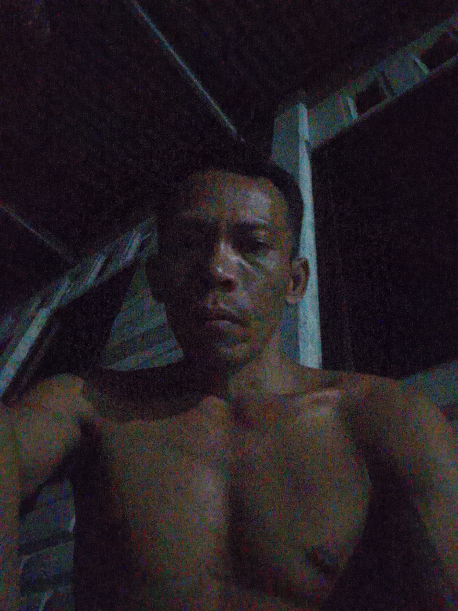 A low-light, grainy, selfie-style photograph depicts a shirtless man with short, dark hair and defined chest muscles. He is positioned against a dimly lit backdrop that appears to be a partially constructed or unfinished building. The background includes wooden beams and a light-colored structural support. The location is likely Bukit Ketok, Indonesia, as specified in the prompt.