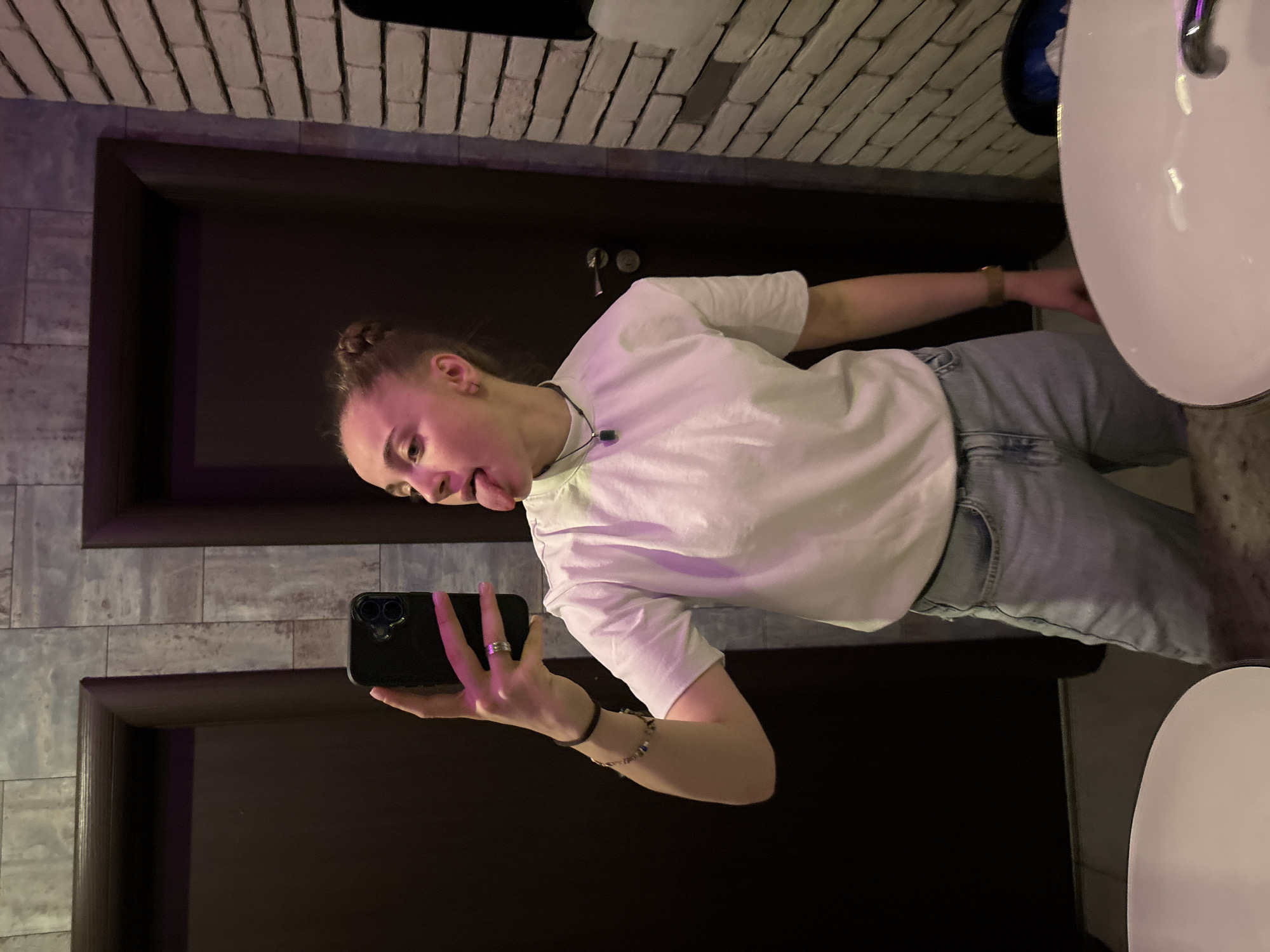 A young woman with a fair complexion, her hair styled in a bun, is seen taking a selfie in a bathroom. She is wearing a white t-shirt and light-wash jeans, and has jewelry on her wrist and fingers. She is sticking her tongue out in the photo. The background includes a dark door with silver hardware, a brick-patterned wall, a gray wall, and a white sink. The image does not provide sufficient evidence to determine the location in Sofia, Bulgaria.