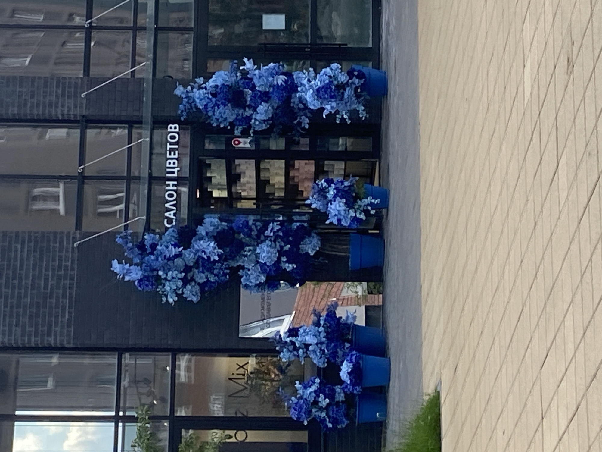 The image shows the entrance to a flower shop, identified by the sign "САЛОН ЦВЕТОВ" (Flower Salon). The shop is decorated with large artificial blue flowers in blue planters. To the left of the entrance, a sign partially visible reads "oz Mix". The facade of the building is dark brick and glass. The street in front of the shop is paved with light-colored tiles. The lighting suggests it is daytime, with sunlight illuminating the scene. There are no people visible in the image. The location is in Yekaterinburg, Russia, specifically at a storefront in an urban setting.