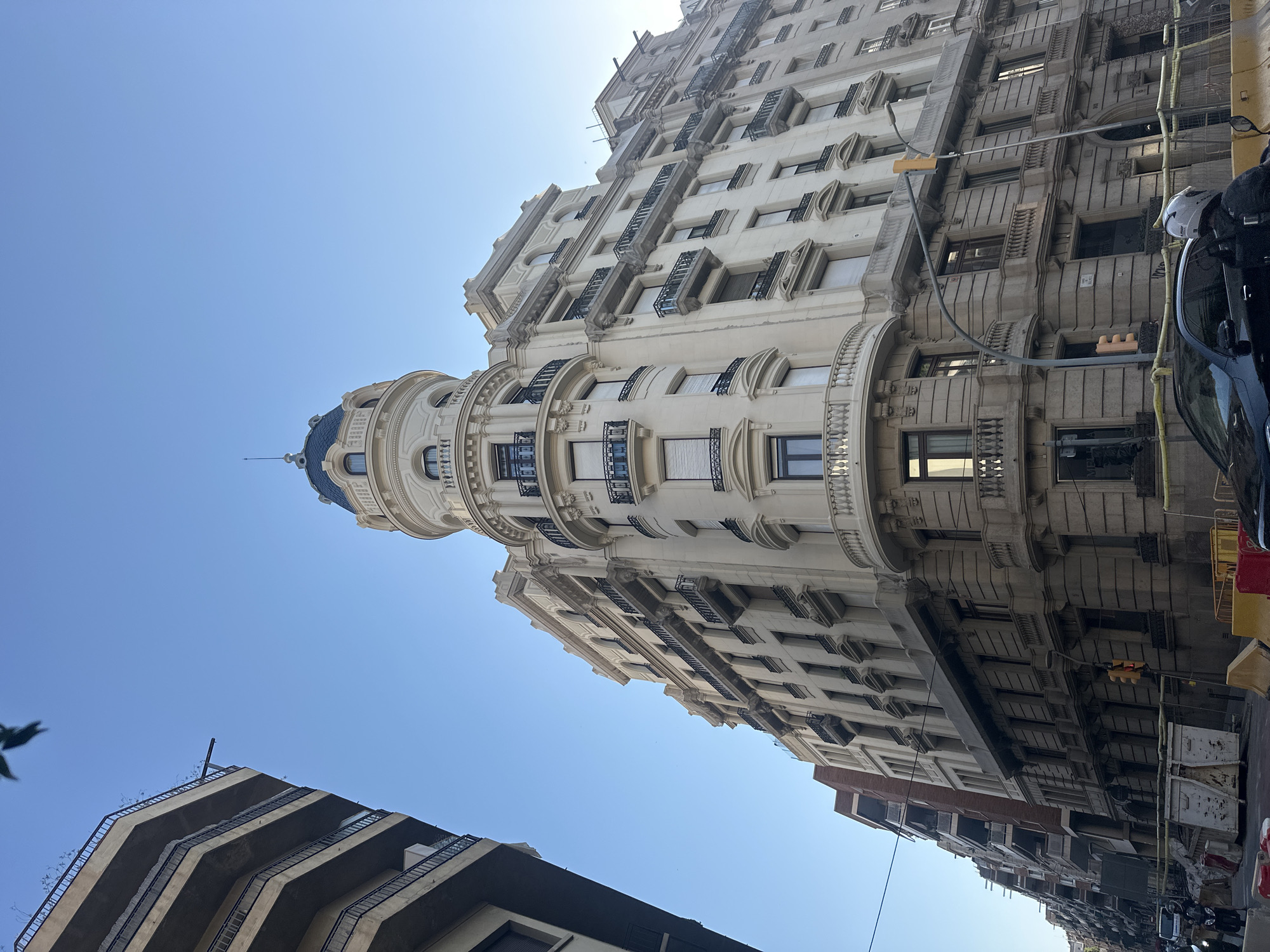 This is a vertical, eye-level shot of a grand, cream-colored building with intricate architectural details, seen from a low angle looking up. The building features multiple stories with numerous windows, many of which are adorned with ornate balconies and shutters. A prominent rounded section of the building culminates in a blue domed roof. The building's facade is decorated with classical elements like columns, moldings, and sculptural reliefs. To the left of the main building, a portion of a more modern, angular building with protruding balconies is visible. The sky is a clear, bright blue, suggesting a sunny day. The scene is captured in a European city, likely Barcelona, given the architectural style and the presence of street elements like traffic lights and parked vehicles on the right side of the frame. The time of day appears to be daytime due to the bright sunlight and clear sky. The overall impression is one of urban architecture, bathed in sunlight.