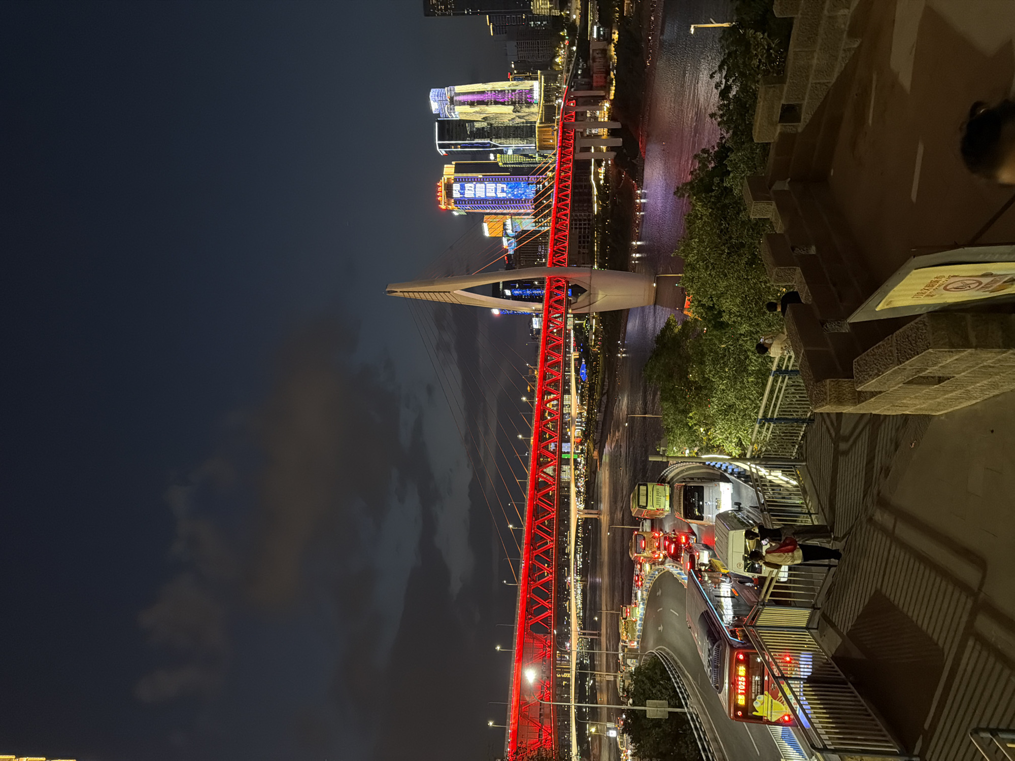 The image captures a nighttime view of a vibrant city scene in Yuzhong District, China, dominated by a brightly illuminated red bridge spanning a body of water. The bridge, a prominent architectural feature, is a cable-stayed bridge with a distinct white pylon. The city skyline, adorned with colorful lights on skyscrapers, is visible in the background.
In the foreground, a busy road curves, filled with traffic. Various vehicles, including buses and cars, are seen navigating the road, their headlights and taillights creating streaks of light. A pedestrian walkway with metal railings overlooks the road and the bridge. Two individuals are standing on this walkway, facing the traffic and the illuminated cityscape.
The environment is characteristic of a bustling urban center at night, with artificial lights reflecting off the water and illuminating the structures. The sky above is dark with some clouds, contributing to the atmospheric nighttime lighting. The overall impression is one of a lively and dynamic city, with a focus on illuminated infrastructure and urban activity.