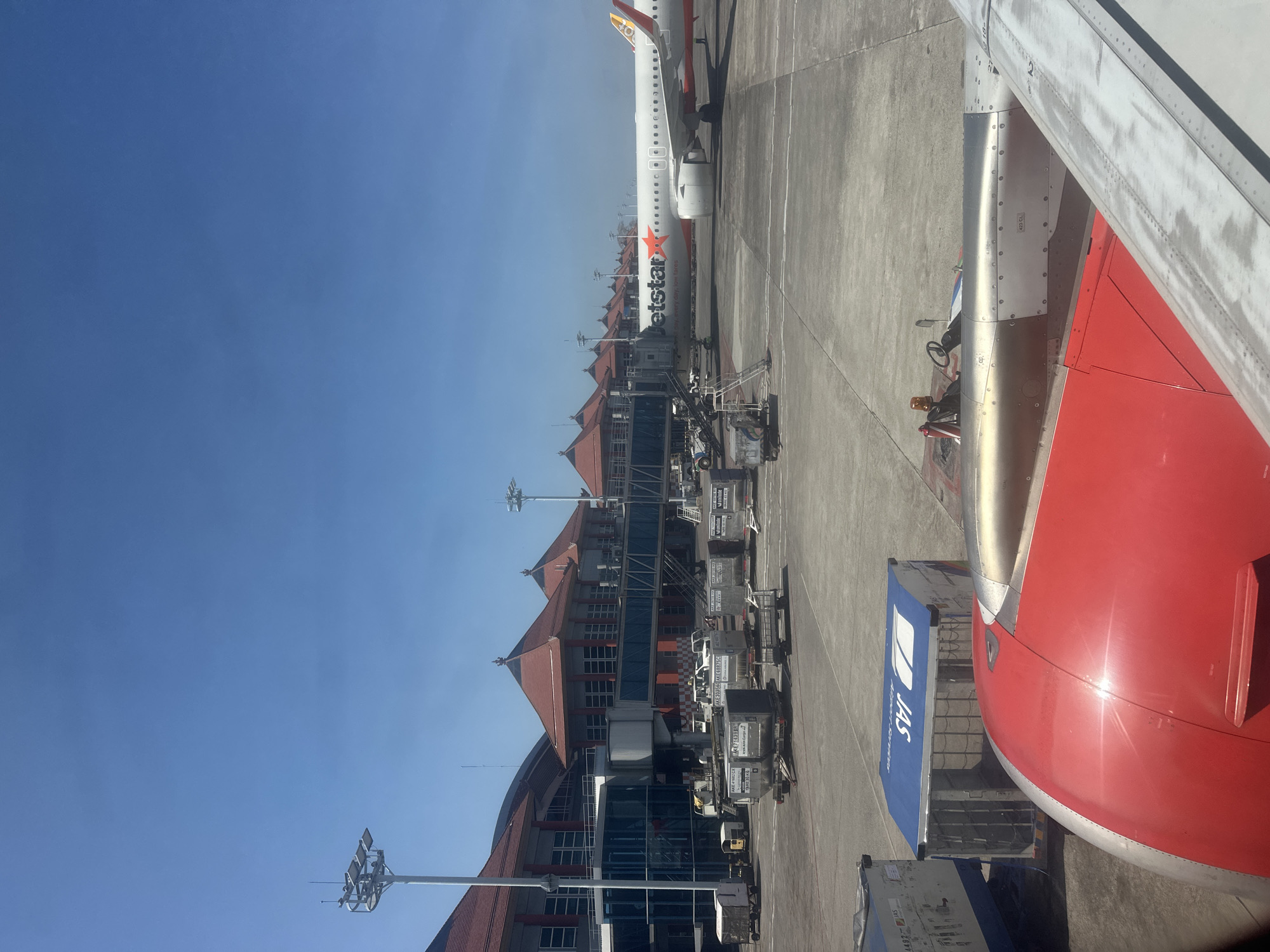 This image depicts an outdoor scene at an airport in Kuta, Indonesia, under a clear blue sky. In the foreground, the wing and red engine cowling of an airplane are visible, suggesting the perspective is from inside another aircraft or at ground level adjacent to an aircraft.
The middle ground of the image shows a Jetstar airplane parked at a gate, connected to a jet bridge. The airplane has the distinctive Jetstar logo and orange star on its fuselage. Beside the airplane and jet bridge, various airport service vehicles and baggage carts are present on the tarmac.
In the background, the airport terminal building is prominent, characterized by its multi-level structure, large windows, and distinctive red-tiled roofs with traditional Indonesian architectural elements. The terminal extends horizontally across the frame.
The overall environment is that of an active airport tarmac on a sunny day, with clear visibility and an emphasis on the aviation infrastructure and equipment.
