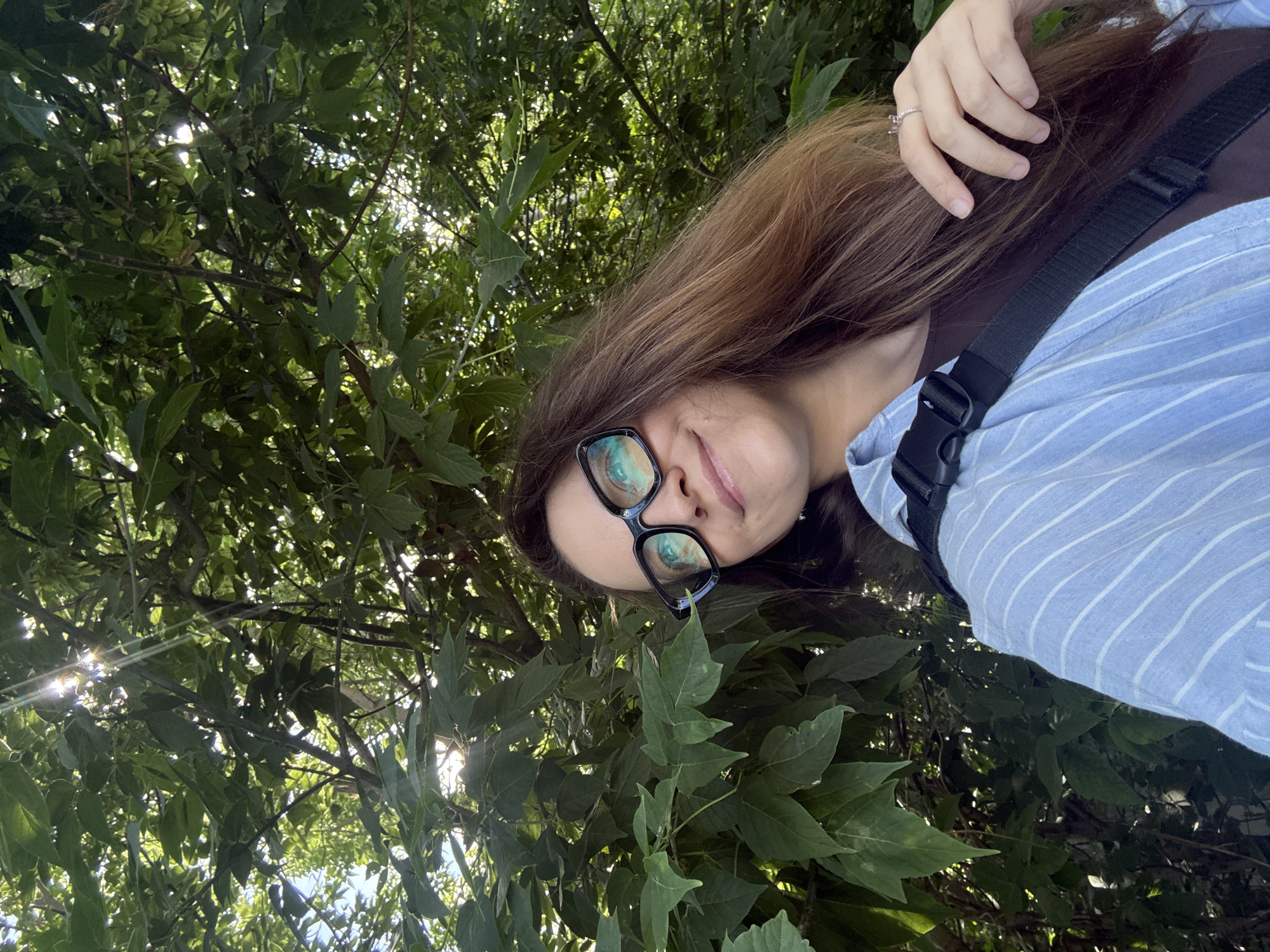 A woman with long brown hair and glasses is taking a selfie outdoors. She is wearing a blue and white striped shirt over a brown shirt, and a black strap is visible across her chest. She is smiling and looking at the camera. Sunlight filters through the green leaves of trees in the background. The location is not specified in the image or provided context.
