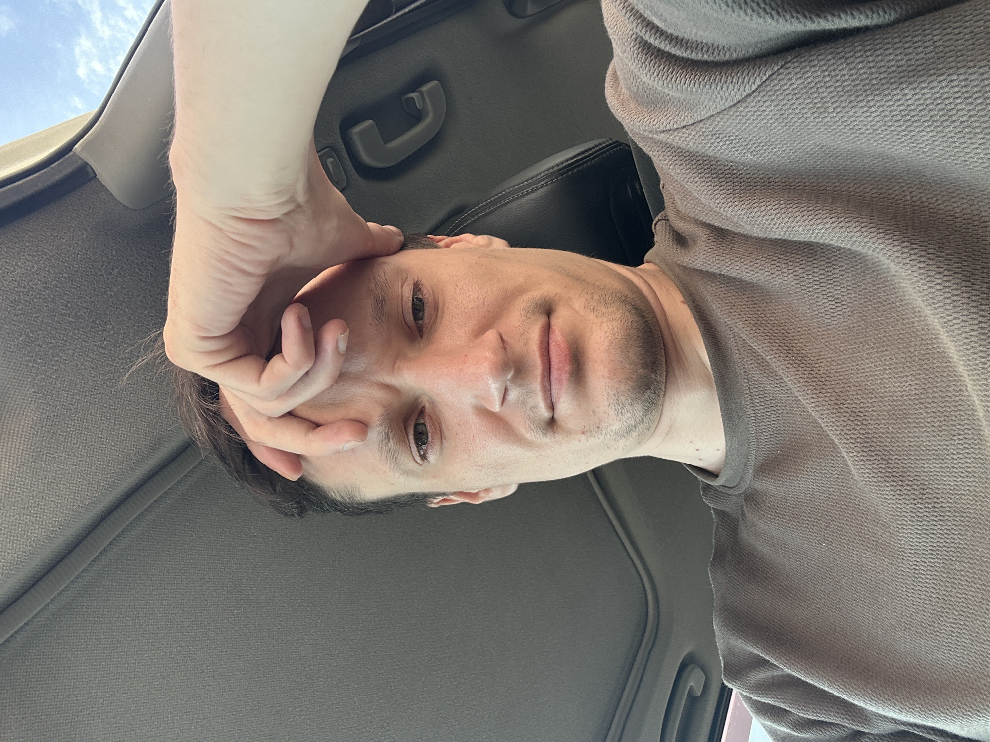 A man is taking a selfie in a car. He has his hand on his forehead, and his eyes are partially closed. He is wearing a brown t-shirt. The car's interior is visible, including the headliner and a grab handle. The sky is visible through the sunroof, indicating it is daytime. The image does not provide information about the city of Shatava, Ukraine.