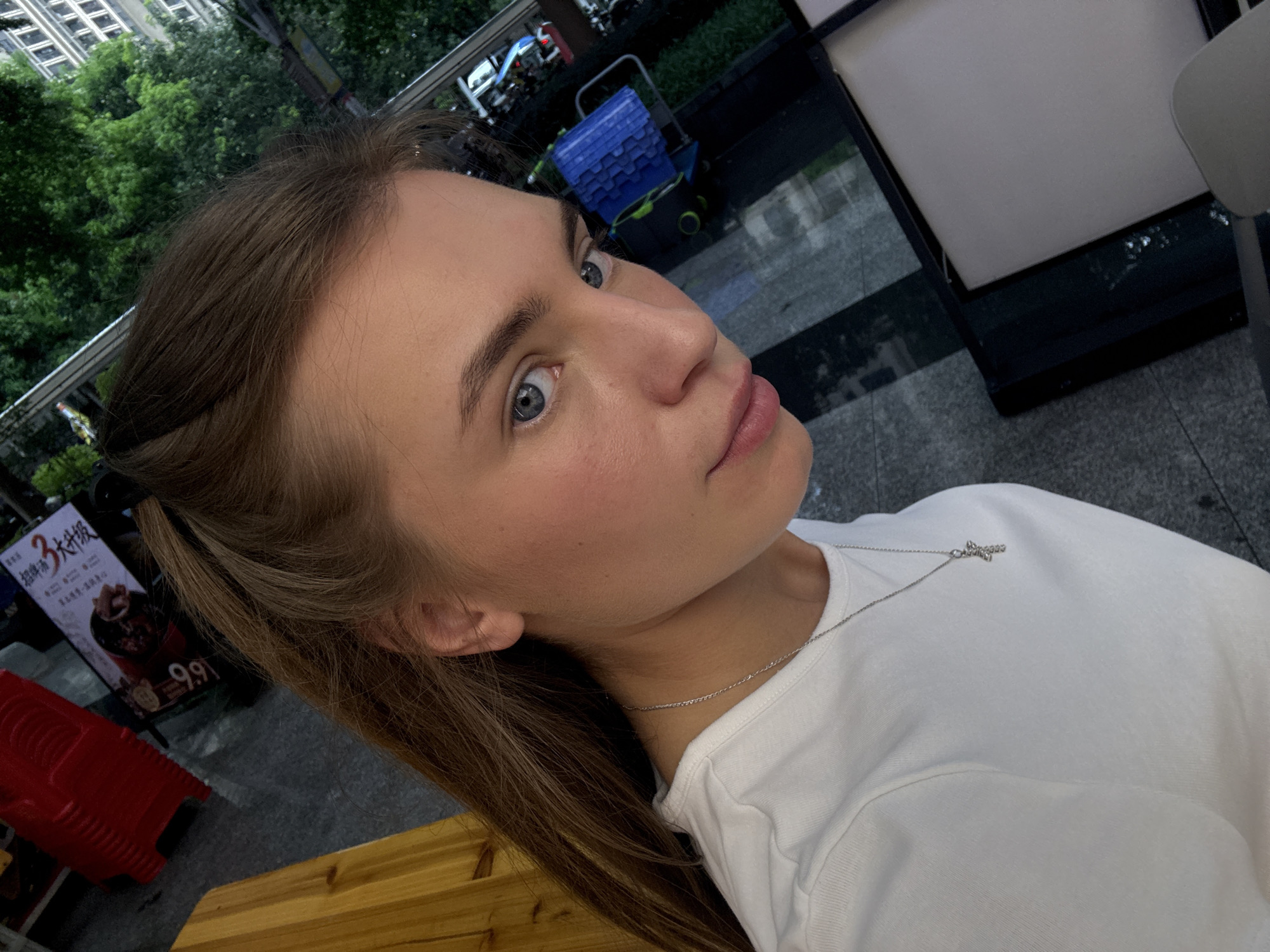 The media file is a close-up, outdoor, eye-level, medium shot of a young woman posing for a selfie. She has long, light brown hair pulled back from her face, with strands framing her head. Her blue eyes are looking slightly away from the camera, and she has a gentle expression on her face. She is wearing a simple white t-shirt and a delicate silver necklace with a cross pendant.
The background suggests an urban environment, possibly a commercial or public area. In the middle ground, there is a blue plastic shopping cart with green accents. Behind it, blurred trees and buildings are visible, indicating an outdoor setting with some greenery and architecture. To the left of the woman, a red object with a ribbed surface is visible, and behind that, a poster with Chinese text and an image of food is partially visible, along with the price "9.9". The lighting suggests it is daytime, possibly late afternoon, with soft, natural light.
The overall mood is casual and relaxed, with the woman appearing at ease. The scene is set in Haizhu District, China, suggesting a contemporary urban backdrop.