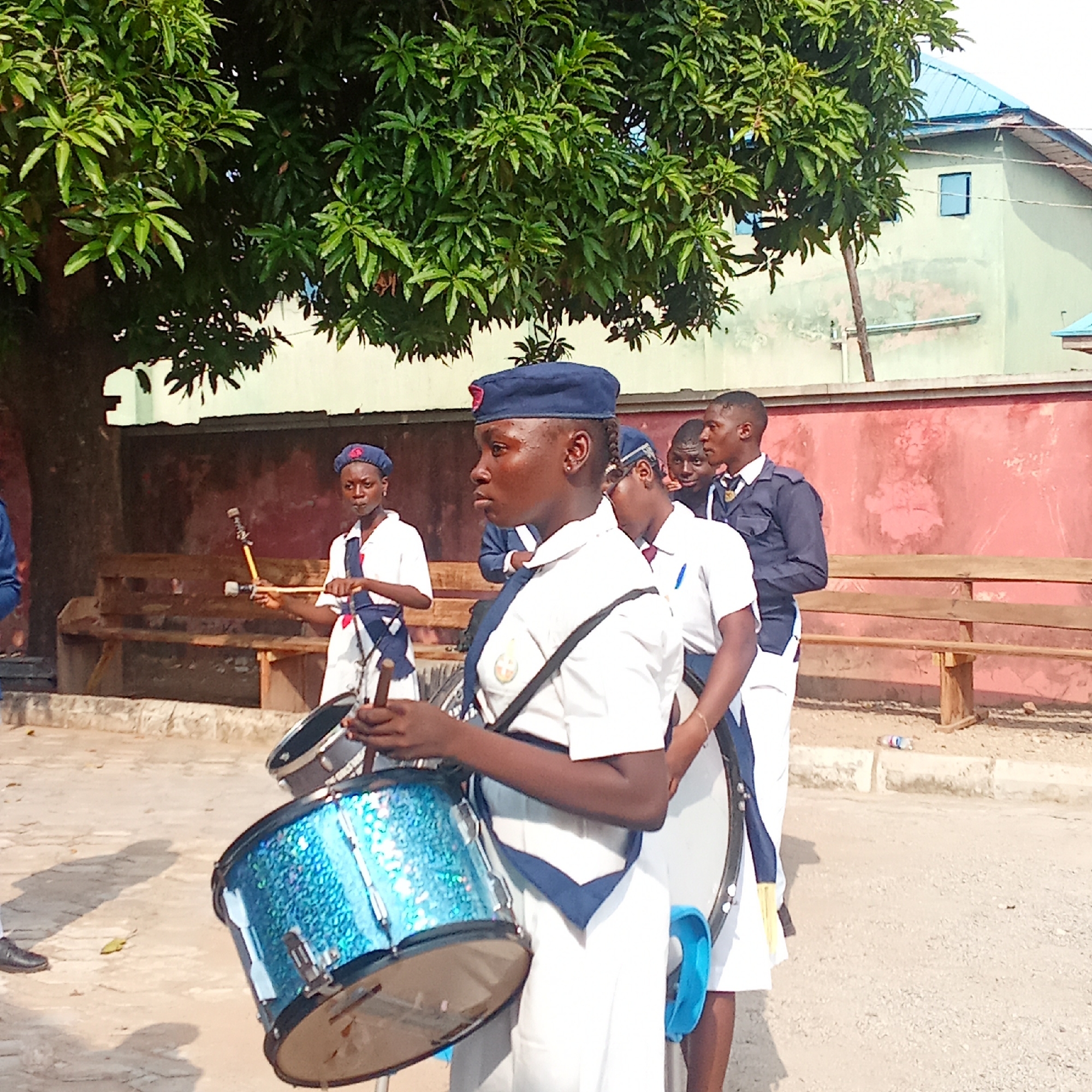 A group of uniformed marching band members are standing in Akwanga, Nigeria. The uniformed members are arranged in a line on a cracked surface. Most members wear white shirts, dark blue hats, and navy blue shoulder straps. One person is carrying a blue drum, and another is carrying a drumstick. A large leafy tree is in the upper part of the image. In the background, there is a red and white wall and a building with a blue roof.