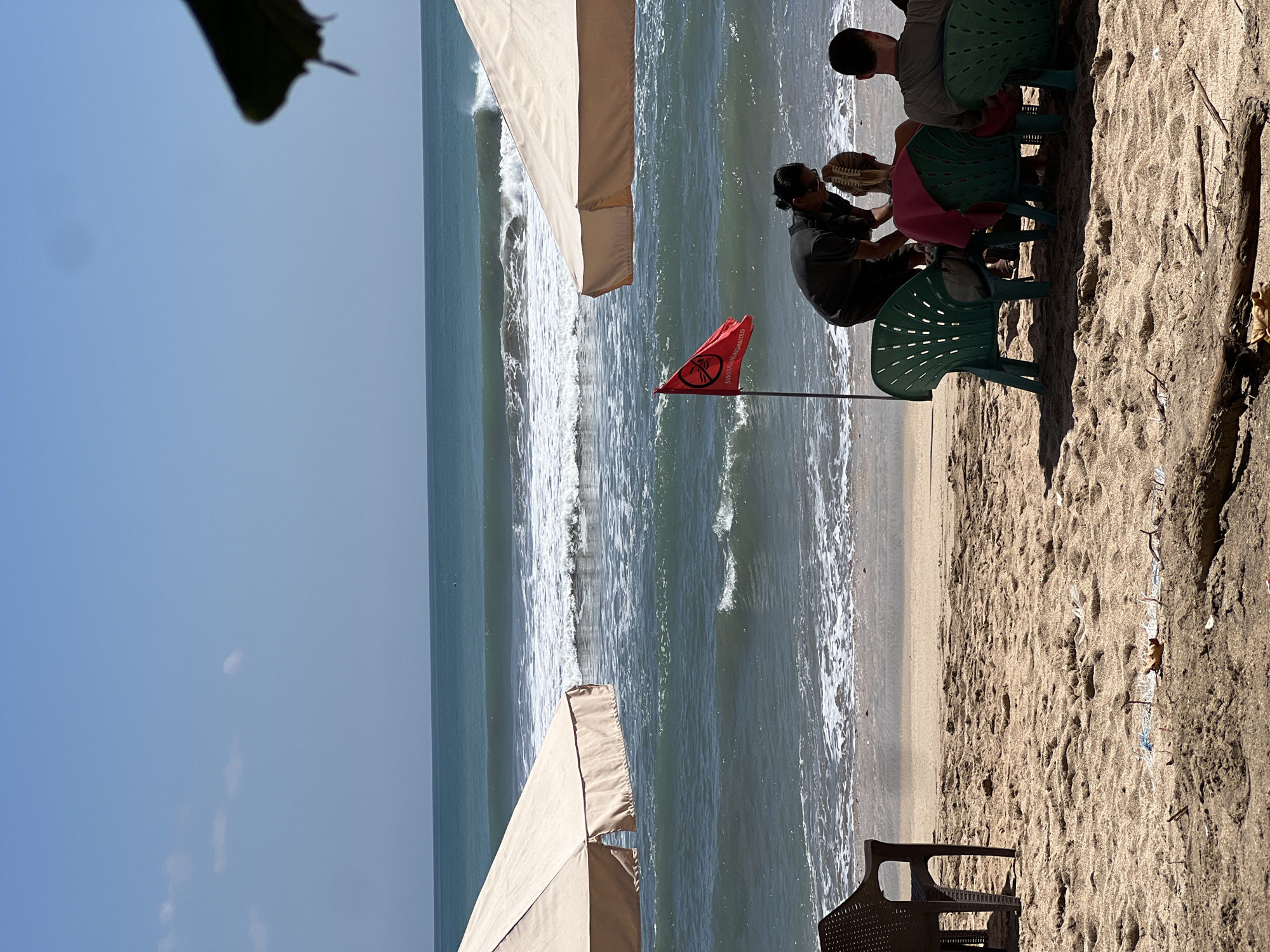 A group of people sit on plastic chairs on a sandy beach in Kuta, Indonesia, under a clear blue sky with a few scattered clouds. The ocean in the background has moderate waves, and a red flag with a "SWIMMING PROHIBITED" symbol is planted in the sand. A person appears to be interacting with another individual sitting in one of the chairs. Two beach umbrellas are visible, one in the foreground on the left and another on the right, partially obscuring the view. The beach itself has some debris scattered on the sand.
