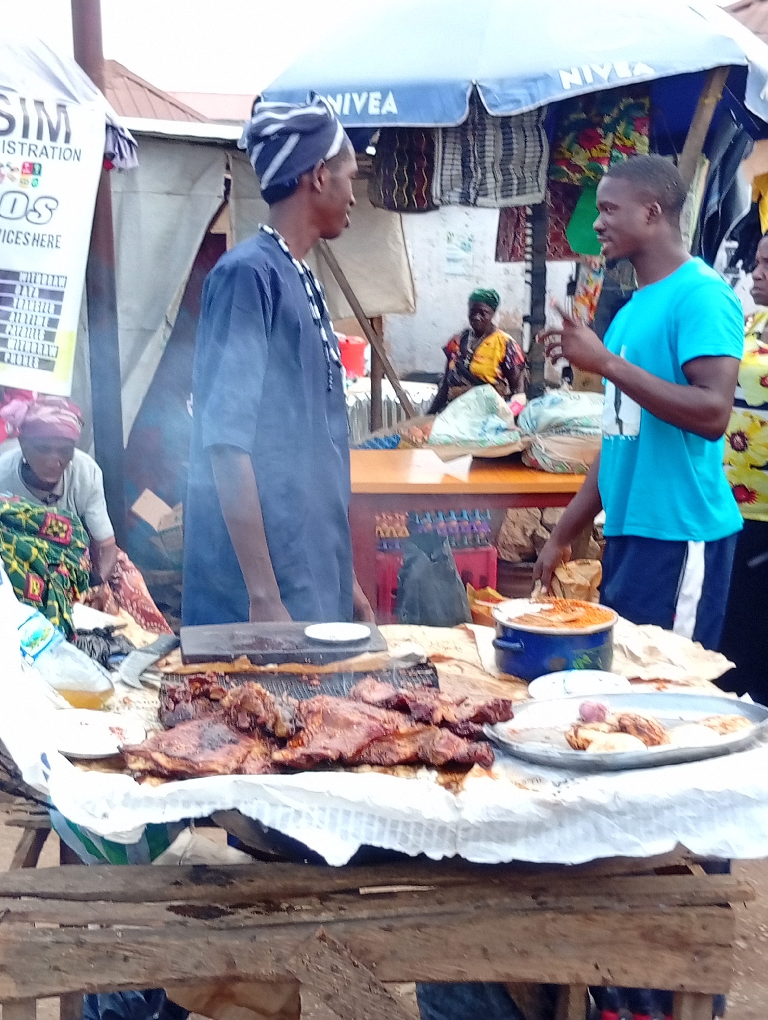 The image depicts a street food stall in Bukuru, Nigeria. A man in a blue t-shirt is seen standing at the stall, facing to the left, and appears to be interacting with a customer or server. In front of him is a platter of grilled meat, and to his right is a blue pot containing food. Behind him, another man wearing a traditional cap and garment stands with his back to the camera. To the left of the frame, a woman in a headscarf and patterned dress is seated, with a bottle of liquid and some raw food items visible on the table in front of her. The stall is set up outdoors, under a blue umbrella that displays the "NIVEA" logo. Various goods are displayed in the background, suggesting a busy marketplace environment.