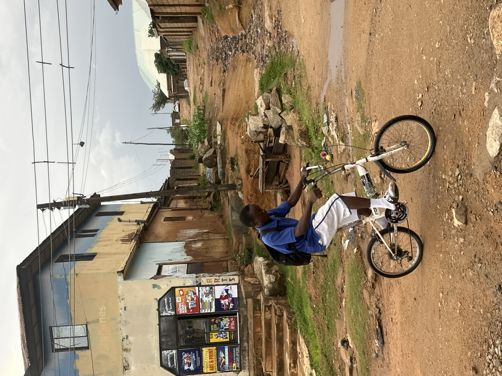 A young Black male is shown riding a bicycle down a dirt road in Gwong, Nigeria. He is wearing a blue t-shirt, white shorts, and white socks. He has a black backpack on. The bicycle is white with a yellow rim on the back wheel. There are two yellow balls on the handlebars. The setting appears to be a village or small town, with various buildings and vegetation. Some buildings are made of brick and some appear to be made of mud or stucco. There is a large, light-colored building with blue and yellow paint on it, and several windows. A shop or store is visible on the ground floor of this building, with advertisements displayed in the windows. One advertisement reads "GRAPHIC DESIGN & PRINT". Another reads "SINISTECH Quality Print". The road is unpaved and appears to have been recently rained on in some areas, with puddles visible. There are piles of rocks and debris along the side of the road. Power lines are visible overhead, strung between wooden poles. The sky is overcast, suggesting it might be a cloudy day. The overall impression is one of everyday life in a rural or semi-rural African community.