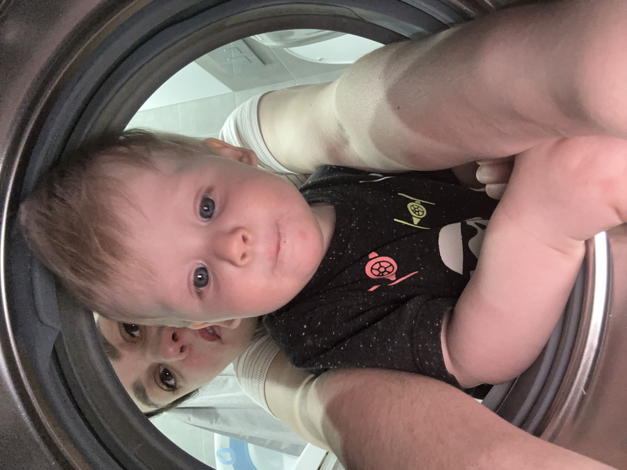 A person, likely an adult, is photographed from inside a stainless steel washing machine with a gray rubber door seal. The face of a small child with fair skin, blue eyes, and light brown hair is directly visible, centered in the frame. Partially visible behind the child is a woman with dark hair and eyes. The adult's arms, with clear tan lines, are visible on either side of the child, suggesting they are holding the child. The child is wearing a dark-colored shirt with a light-colored Star Wars logo on the right side of their chest. The background visible through the washing machine door is blurry, but tile and what appears to be bathroom fixtures suggest the location is likely a bathroom. There is no visual indication that the image was taken in the city of Chaikovsky, Russia. The interaction is an adult holding a child while being photographed inside a washing machine.
