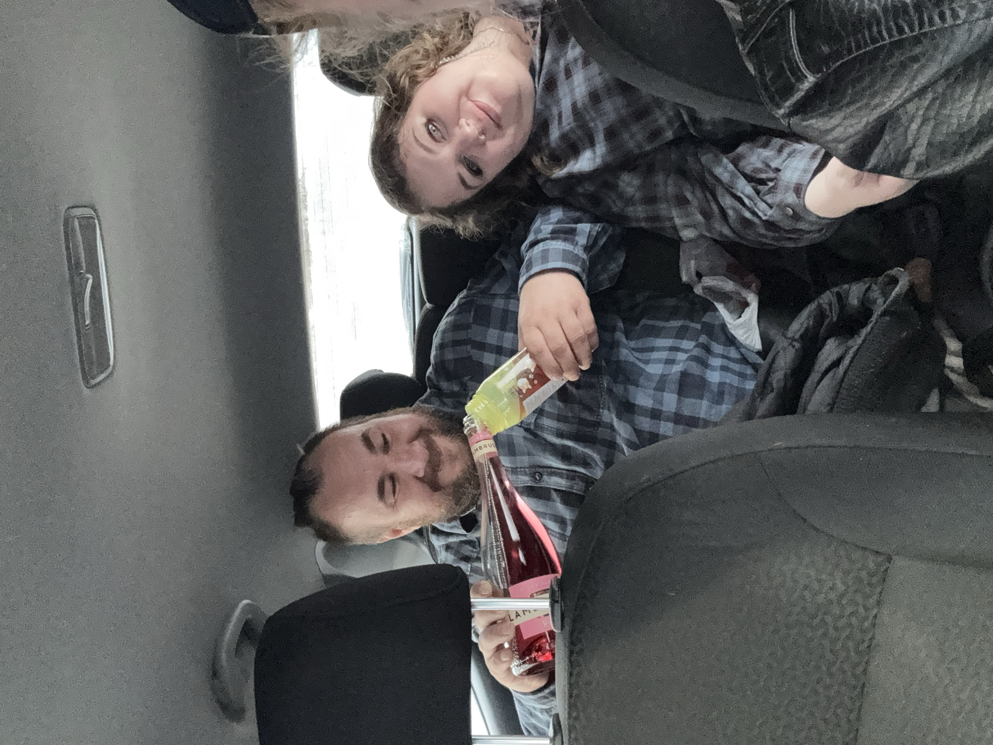 Two people are in the back seat of a car. A bearded man is pouring pink liquid from a bottle labeled "LAMBRUSCO" into a glass. A woman is sitting next to him, facing the camera. The interior of the car is visible, including the car door and seat. The city of Izhevsk, Russia is not apparent in the image.
