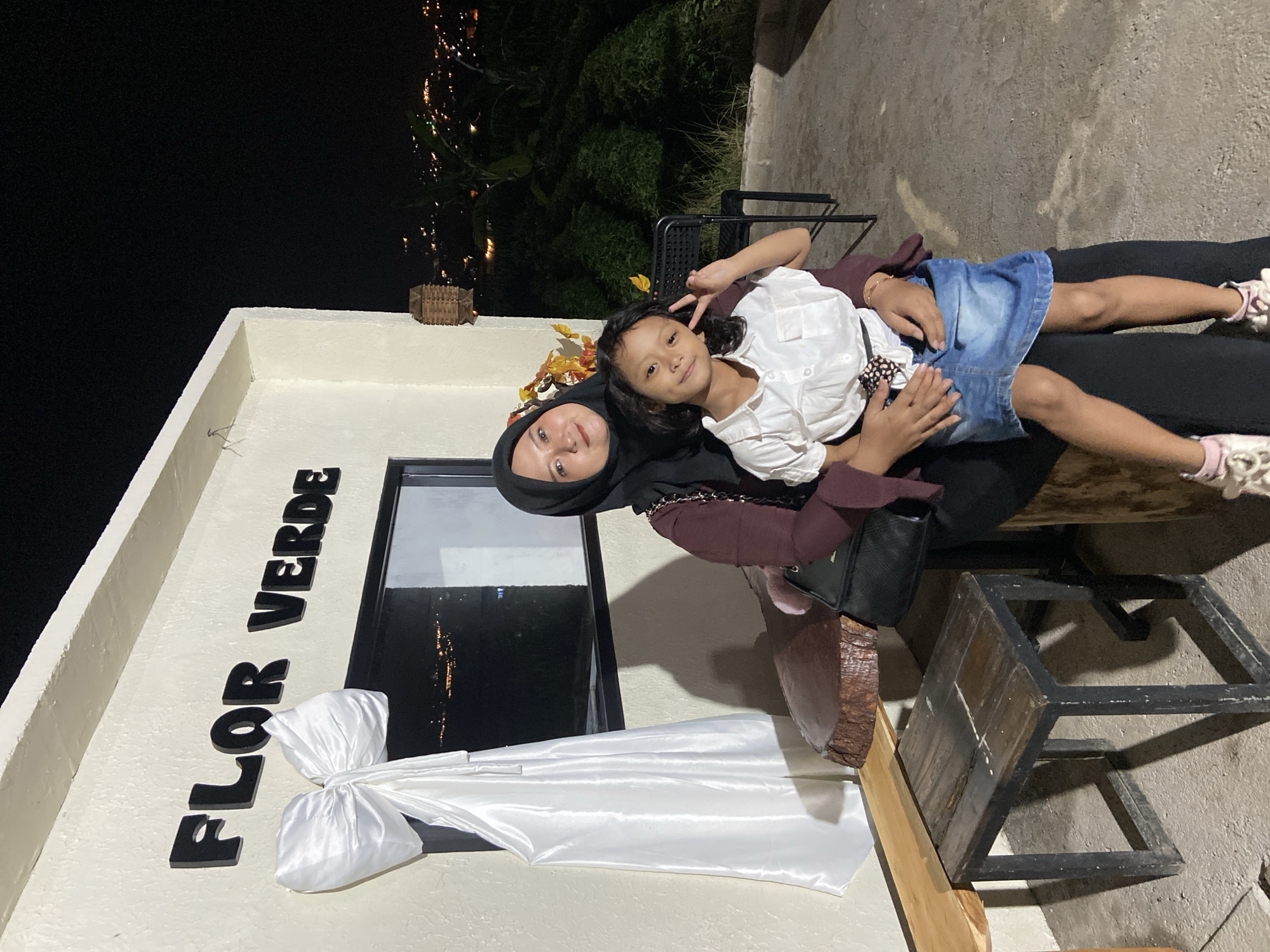 The image shows a woman and a young girl posing for a picture outdoors at night. The woman is wearing a black hijab and a maroon shirt, while the girl is wearing a white shirt and a denim skirt. The girl is lying on the woman's lap, with her arm raised in a peace sign. The background shows a building with the words "FLOR VERDE" in large black letters. There is also a window with a reflection of lights, suggesting a city view at night. The overall mood appears to be happy and relaxed. The setting seems to be a patio or outdoor seating area, possibly a restaurant or cafe, given the seating and decorative elements. The time of day is clearly night, as indicated by the darkness and the lights in the background. The weather appears to be clear. The location context provided is Kesiman, Indonesia.