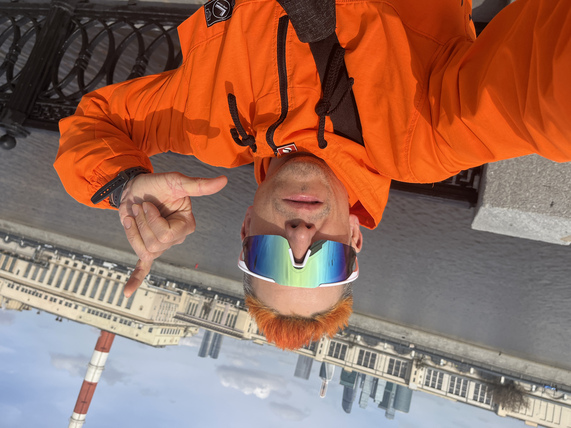 A selfie taken in Moscow, Russia shows a fair-skinned person wearing an orange jacket and sunglasses with mirrored lenses. The individual points downward with their right index finger. The person has orange hair and a light beard. In the background are buildings, including the Moscow City skyscrapers. The Moskva River and embankment are also visible. A red and white smokestack rises in the distance.