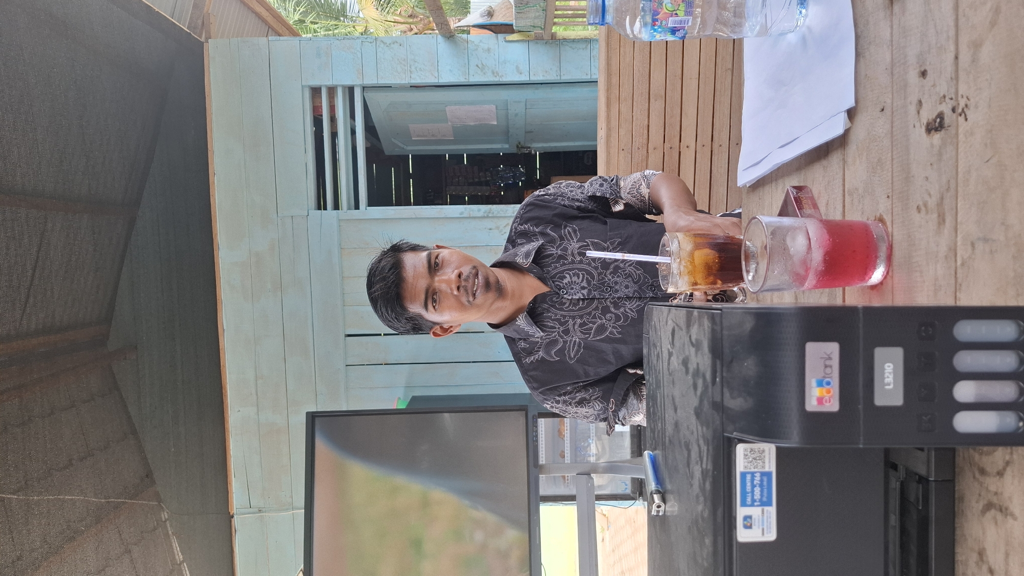 A man with dark hair and a mustache, wearing a black collared shirt with a white patterned design, is seated at a wooden table. He is looking towards the camera. In front of him are two glasses with drinks, one with ice and a straw, and a bottle of water. To his left is a black printer with an "ecotank" logo and a model number "L3210". A screen or monitor is also visible to his left. The background shows a light blue wooden structure, possibly a building or shelter, with an open doorway. Wooden planks form seating or a counter area behind the table. The environment appears to be outdoors, with vegetation visible in the background. The scene is set in Grong-grong, Indonesia.