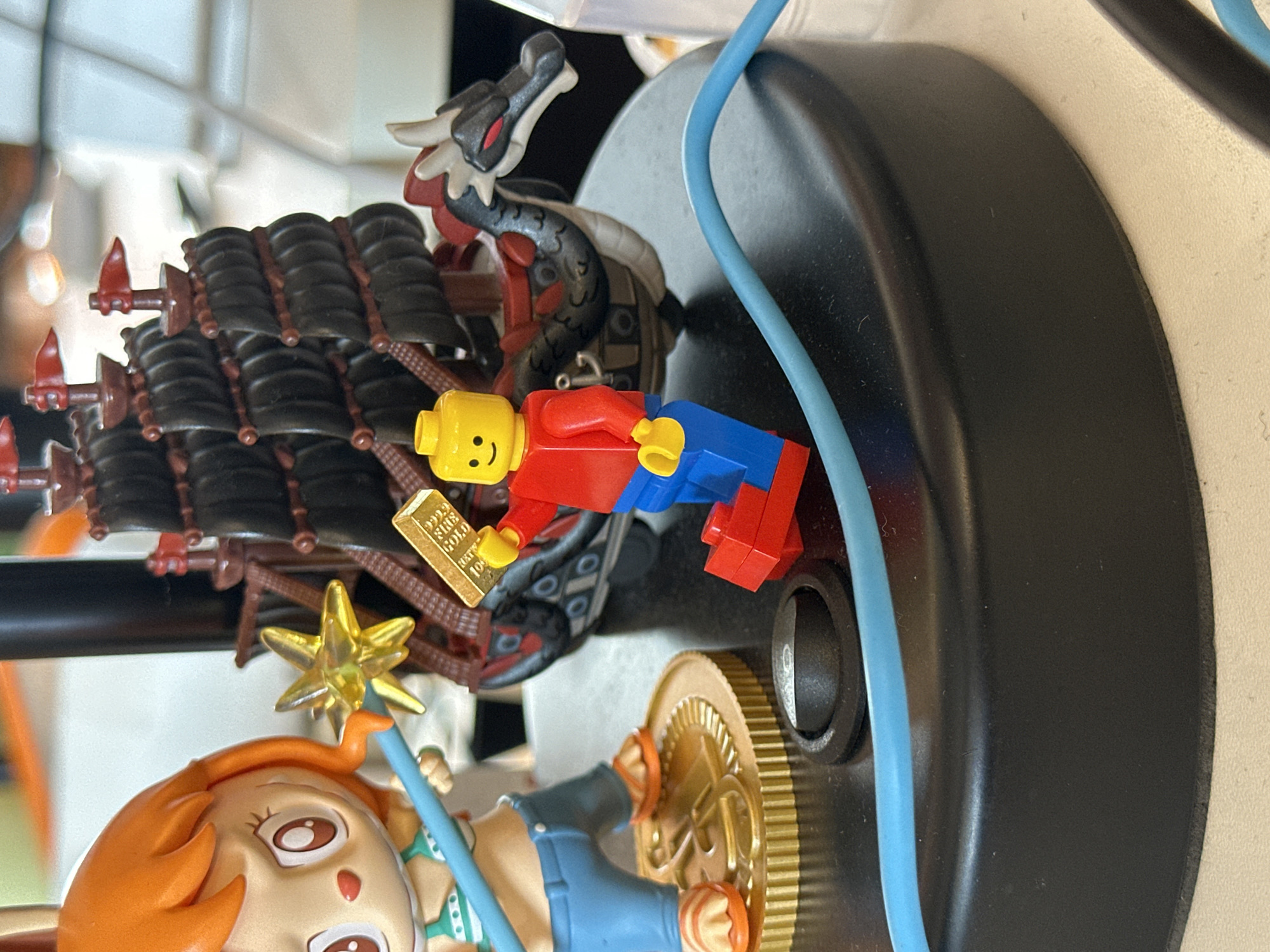 The image depicts a collection of toys and figurines. A Lego minifigure with a red torso, blue legs, and a yellow head is holding a gold bar. Behind the minifigure is a toy dragon with a grey body and red and white accents, along with a section of what appears to be a ship with black sails. In the foreground is an orange-haired figurine holding a star-shaped wand. These objects are situated on a black, rounded base with a gold coin. A blue cord is present near the base. The image does not provide information about the location in the city of London, United Kingdom.