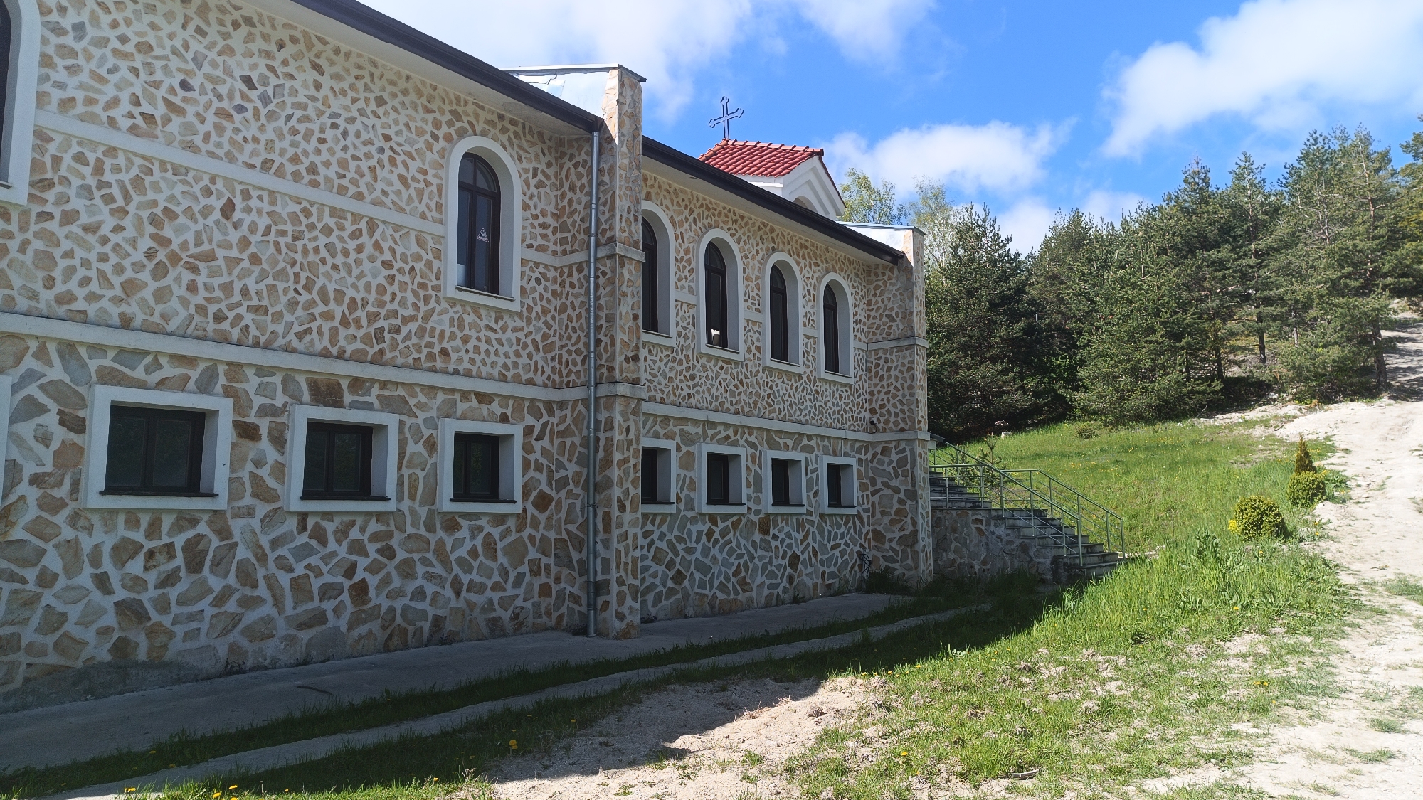 The image shows the exterior of a stone building in Sini Vrah, Bulgaria. The building has two stories with rectangular windows on the first floor and arched windows on the second floor. The walls are constructed of irregular shaped stones. A red-roofed structure with a cross on top is visible behind the main building. The surrounding landscape includes a grassy area with a dirt path and pine trees. A set of stone stairs with a metal railing leads up a grassy hill. The sky is blue with scattered white clouds.