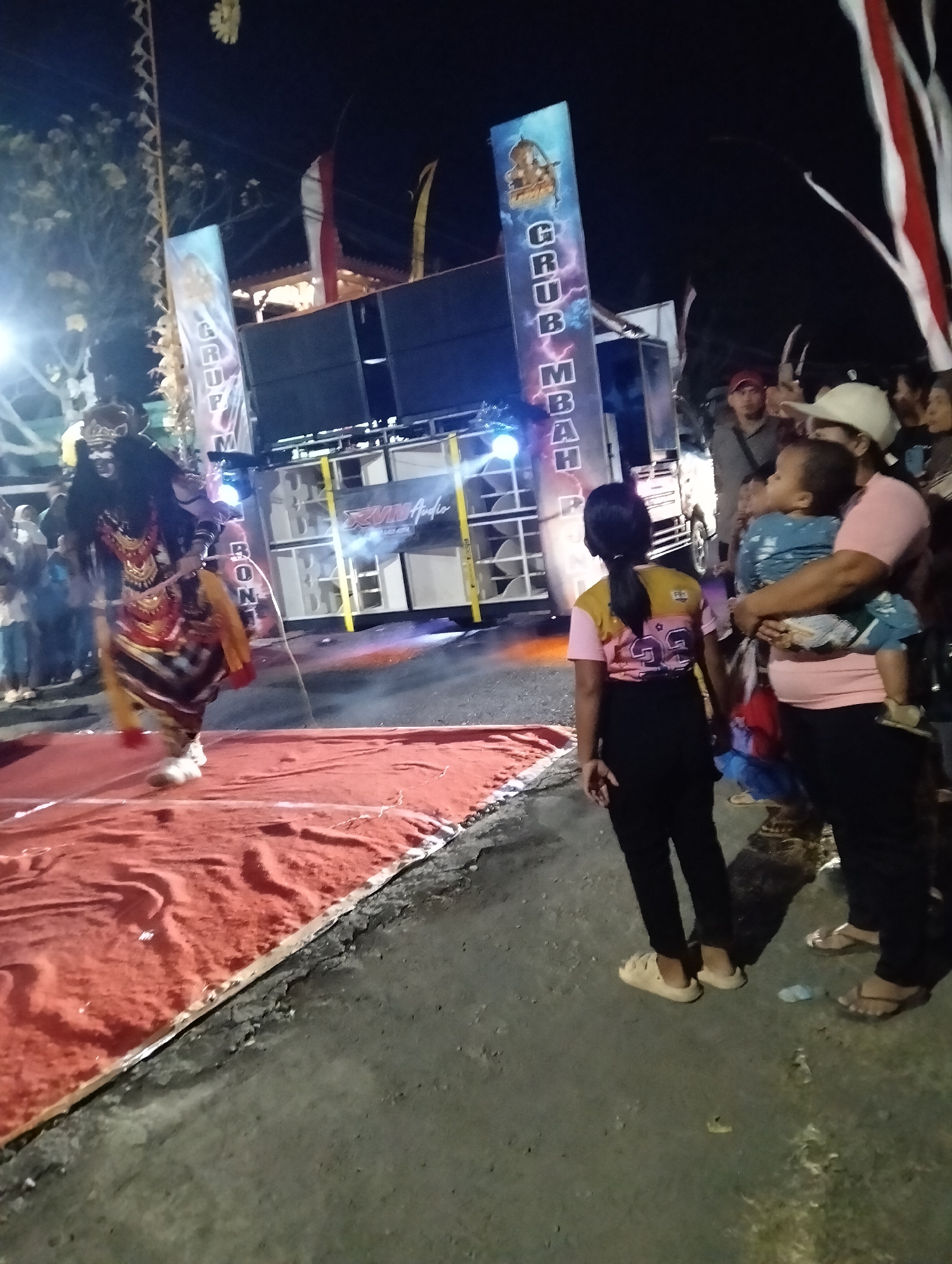 The image captures a nighttime outdoor scene, likely a performance or event, in Sumbersewu, Indonesia. In the foreground, a performer is dressed in a traditional, elaborate costume featuring a dark headdress and colorful, patterned garments, standing on a red carpet. To the right, a woman is holding a baby, both facing the performance. Behind them, a large speaker system is set up, with a banner displaying vertical text, including "GRUMB" and "MBAR". A group of people are gathered in the background, some partially obscured. The environment appears to be a street or open area, with visible lighting and dark, rough asphalt ground. The overall atmosphere suggests a lively cultural event.