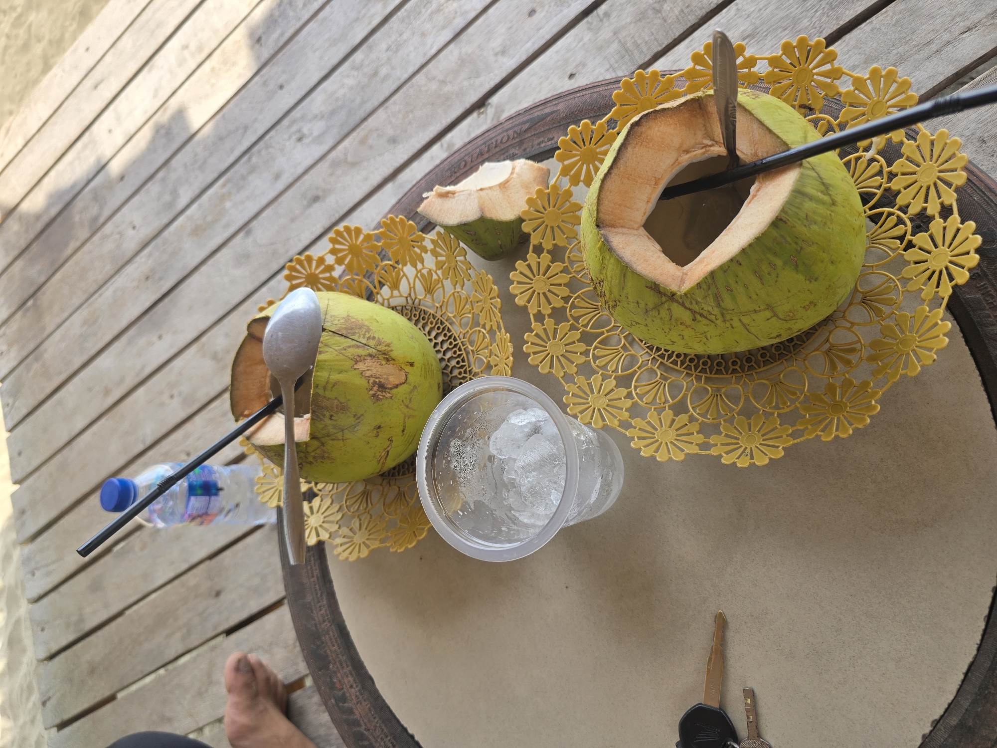 On a light-colored wooden table, two green coconuts are prepared for consumption. Each coconut has been partially opened and a black straw inserted. One coconut is positioned to the left and slightly further away, with a spoon resting on its rim. The other coconut is closer to the foreground and to the right, also with a straw. Next to the coconuts, a clear plastic cup is filled with ice cubes. A bottle of water with a blue cap and a straw is visible behind the upper coconut. In the lower left corner, a set of keys lies on a brown surface. A fragment of a coconut, possibly a husk or shell, is situated to the right of the upper coconut. The table is set on a wooden deck, with what appears to be sand visible in the upper left background, suggesting an outdoor setting. The lighting suggests it is daytime. The scene is set in the city of Reudeuep, Indonesia.