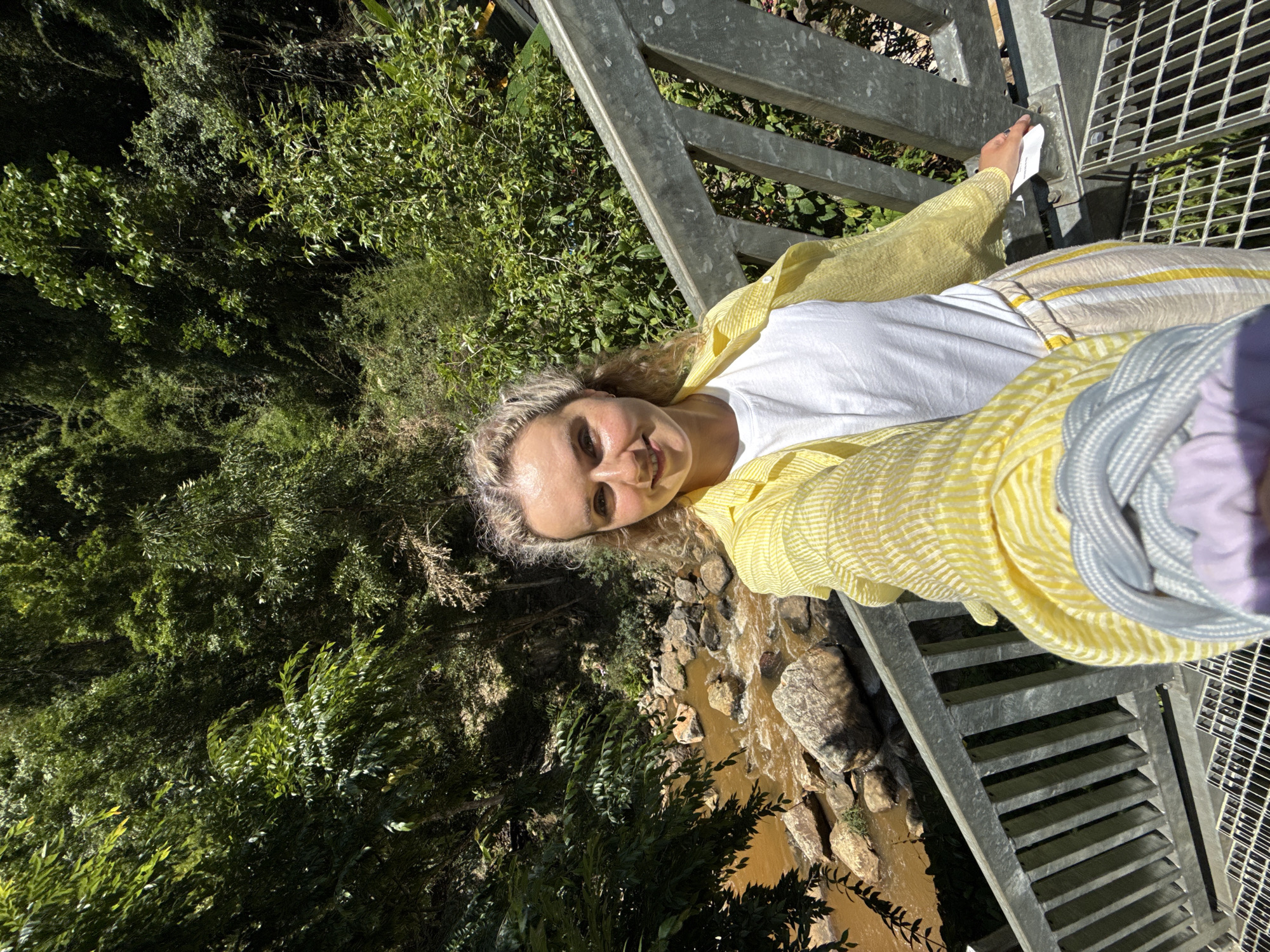 A blonde woman is positioned on a gray metal staircase or bridge. She is wearing a white t-shirt and a yellow and white striped shirt/jacket. The orientation suggests she may be holding a device to capture the image as a selfie. In the background, there's lush green vegetation to the left and below the bridge, along with a water stream or river with visible rocks. The water appears to be a muddy brown color. The metal structure and surrounding landscape suggest an outdoor environment, possibly a park or natural area. Given the request for a location, there is no confirmation that this image was taken in Da Lat, Vietnam, unless explicitly given as a location in the image itself.