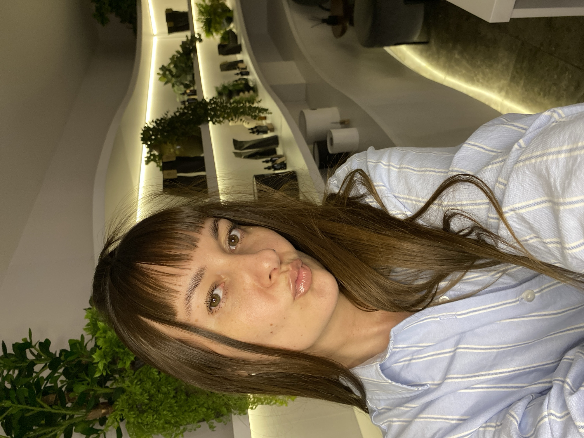 A medium shot depicts a woman with brown hair and bangs, wearing a light blue striped shirt. She is posed with pursed lips. In the background, there is a curved wall display featuring various plants and objects. The setting appears to be an indoor space. The location is not specified to be in Votkinsk, Russia.