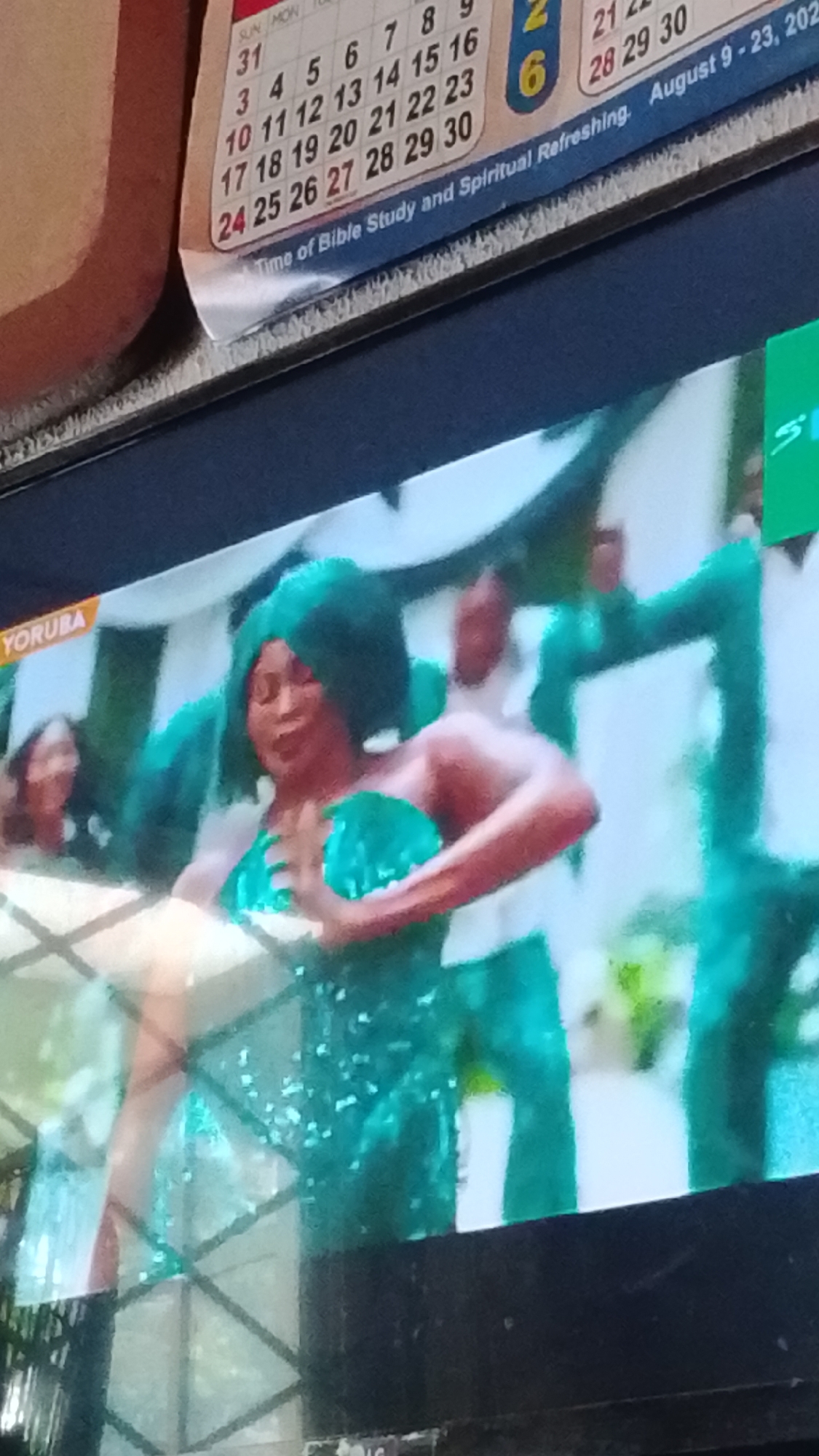 A television screen displays a scene featuring a woman in a sparkly green outfit and afro-style wig, with other individuals in similar green attire behind her. A yellow "YORUBA" label is visible in the upper left corner of the screen, suggesting the program is related to Yoruba culture. In the background, a calendar is partially visible with dates for August 2023. The calendar shows "Time of Bible Study and Spiritual Refreshing. August 9 - 23, 2023." This indicates the program is likely being aired in Nigeria, potentially Lokoja, as indicated by the request, although the image itself does not explicitly confirm the location within the city.