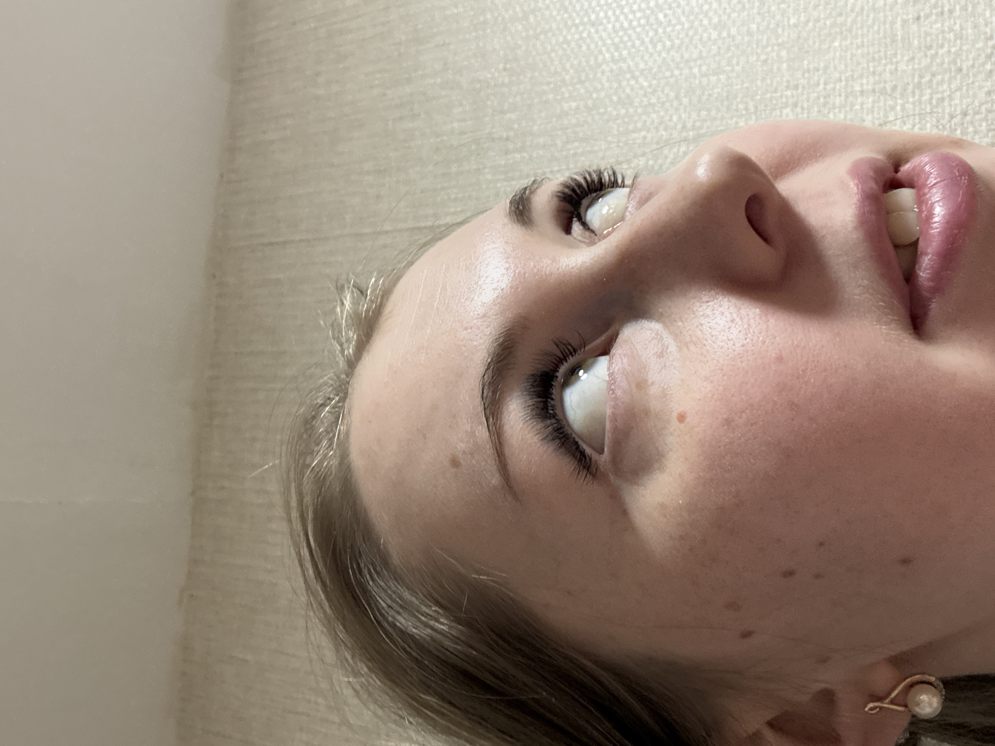 A close-up portrait, oriented sideways, features the upper body and face of a young adult woman. She is positioned horizontally in the frame, with her head tilted back and looking upwards. Her light skin is visible, with hints of light-colored hair (blonde or light brown) near her temples. Her eyes are open wide, revealing prominent, long, and dark eyelashes. Her dark eyebrows are well-defined. Her mouth is slightly open, showcasing white teeth and full, glossy pink lips. Small moles or freckles are visible on her forehead and along her cheek/neckline. A pearl earring with a gold-colored wire is seen in her right earlobe. The background consists of a light-colored, textured surface, indicating an indoor setting. The lighting is bright and even, but the specific time of day is not apparent. The woman's expression appears neutral or contemplative. There is no visible text in the image.