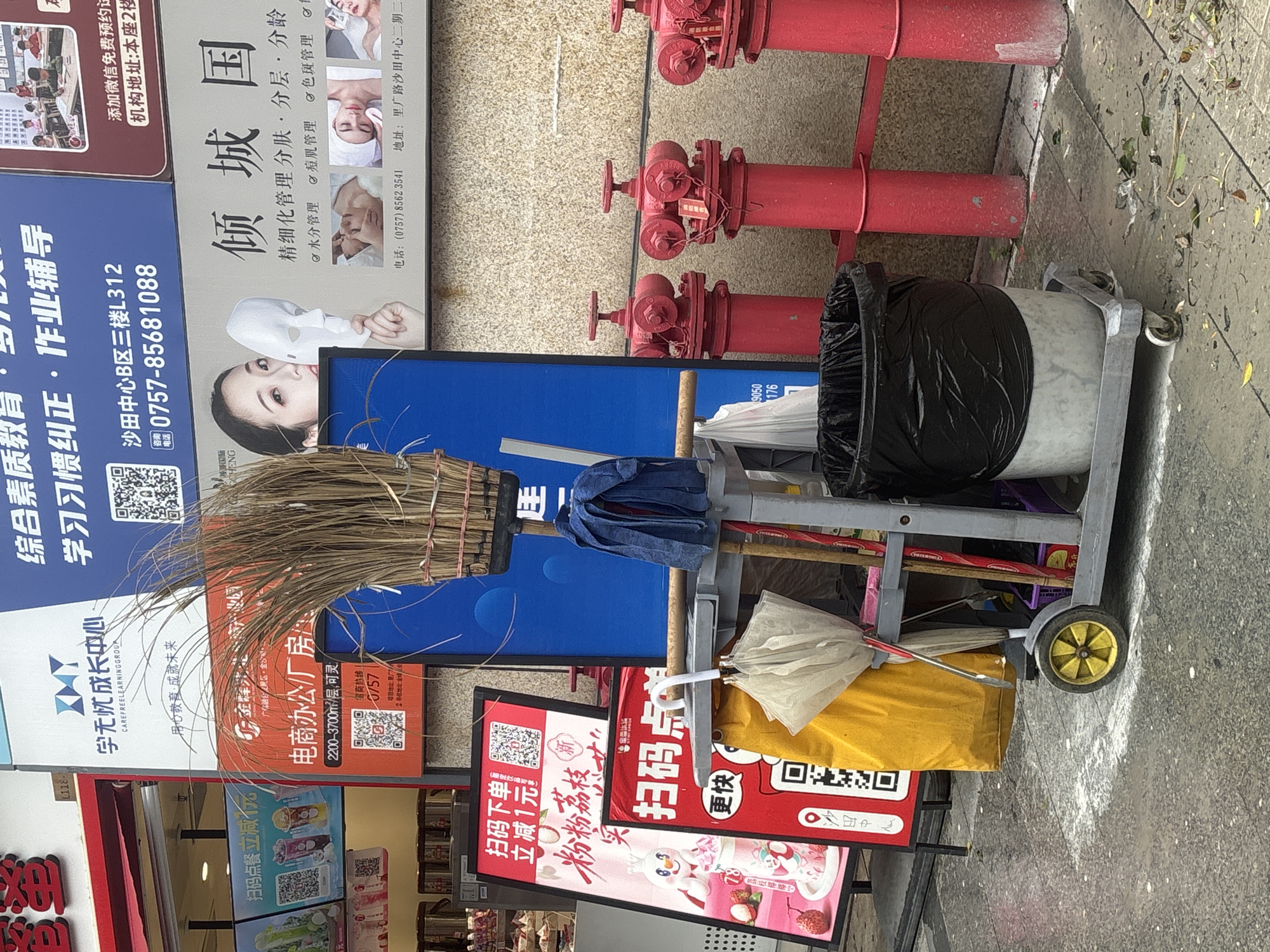 The image captures a street scene in Nanhai District, China, likely in a commercial area. In the foreground, a cleaning cart stands on a wet, paved surface. The cart holds a broom with dried reeds, a blue mop, a yellow bag, a rolled-up umbrella, and a black trash bag. Various signs are visible in the background, including advertisements for a learning center, a beauty salon, and a food establishment. The building facades are adorned with commercial signage, and several red fire hydrants are present along the wall. The environment appears to be outdoors, with the ground showing signs of recent moisture or spillage.
