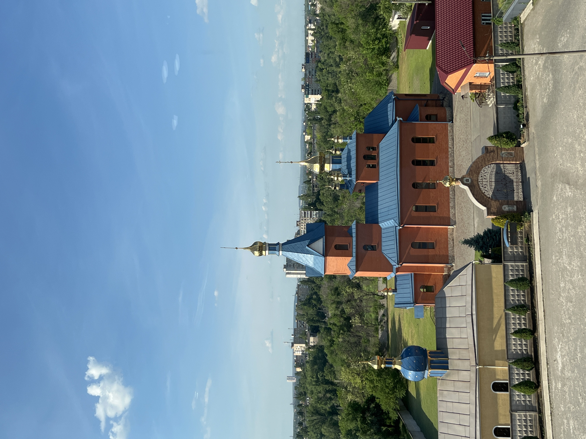 This is an elevated view of a church complex located in Kryvyi Rih, Ukraine. The building features orange facades, blue roofing, and golden domes, and it is bordered by green trees and residential buildings. A paved road runs along the side. The sky is a clear blue with few clouds. The complex is accessible through an ornate gate.