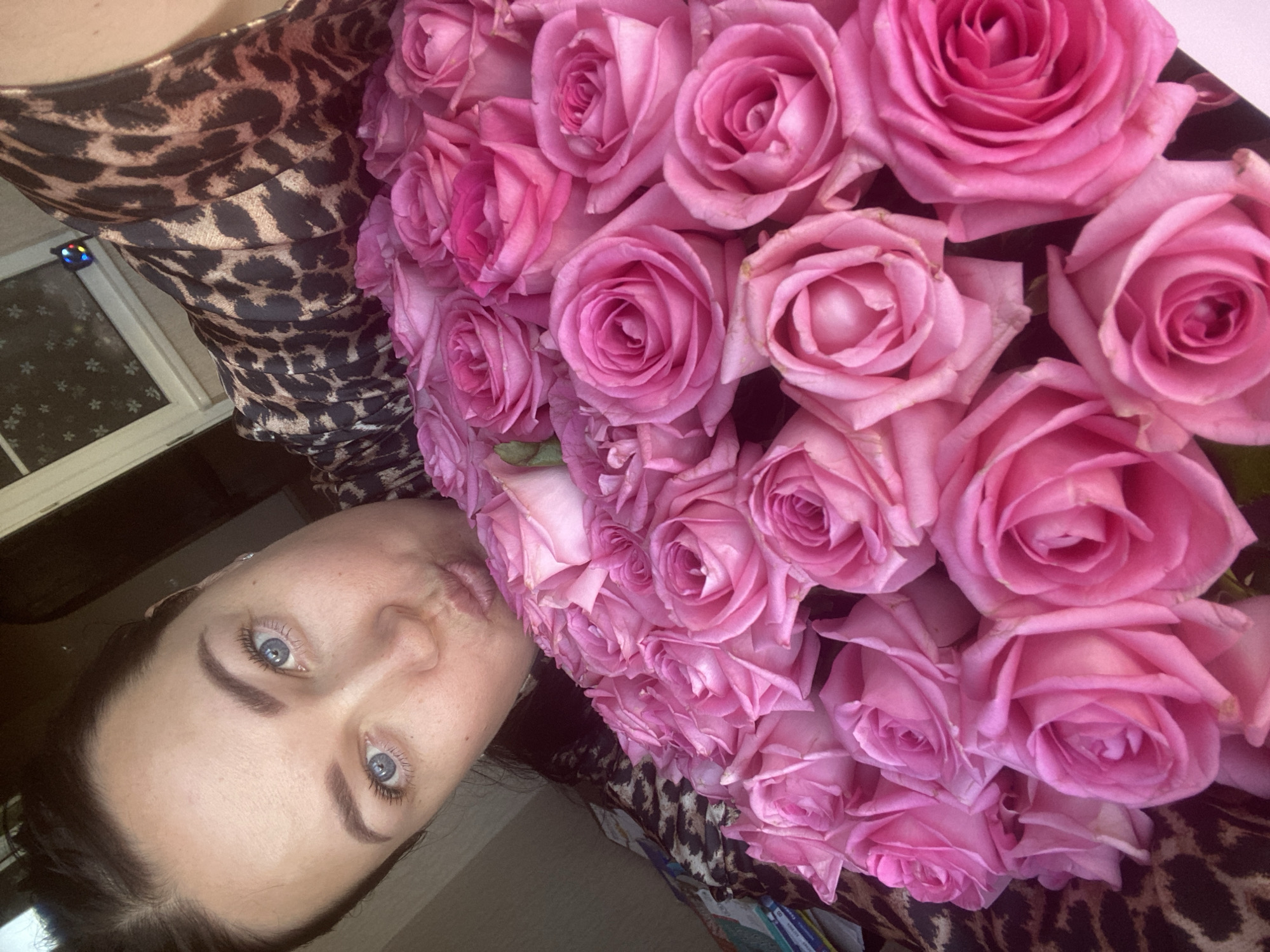 A woman with fair skin and blue eyes is holding a large bouquet of pink roses. She is wearing a leopard-print dress. The background shows a window with a visible reflection of a nearby building. Based on the mention of Sochi, Russia, the location is inferred to be somewhere in that city.