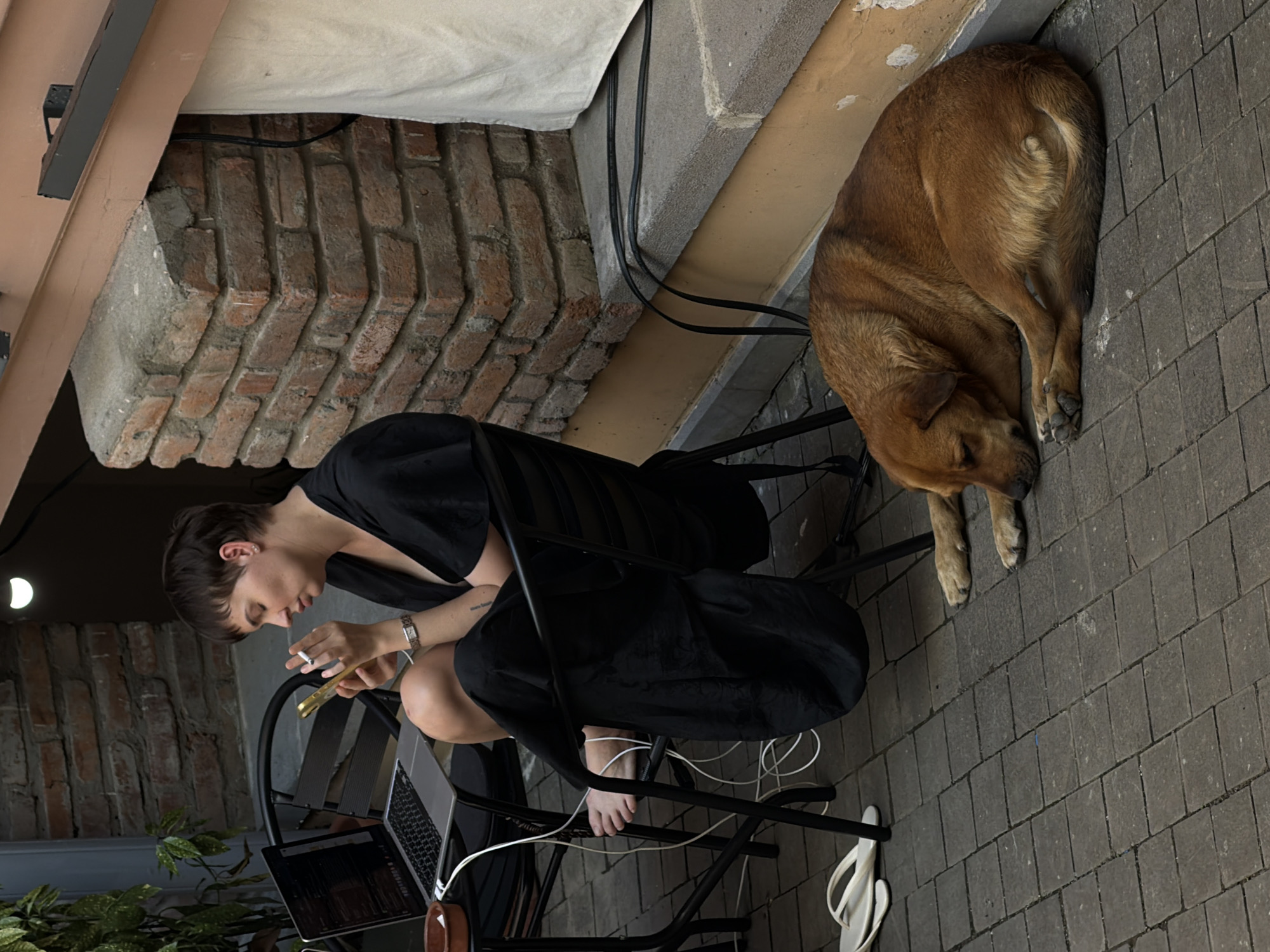 The image shows a woman sitting on a chair outdoors, looking at her phone and smoking. A laptop is on a small table next to her, and a dog is sleeping on the ground in front of her. The scene appears to be in an urban setting, possibly a courtyard or patio area, with brickwork visible in the background. The lighting suggests it is daytime, but possibly overcast or late in the day. This scene could be located in Tbilisi, Georgia, a city known for its eclectic architecture and outdoor cafe culture.