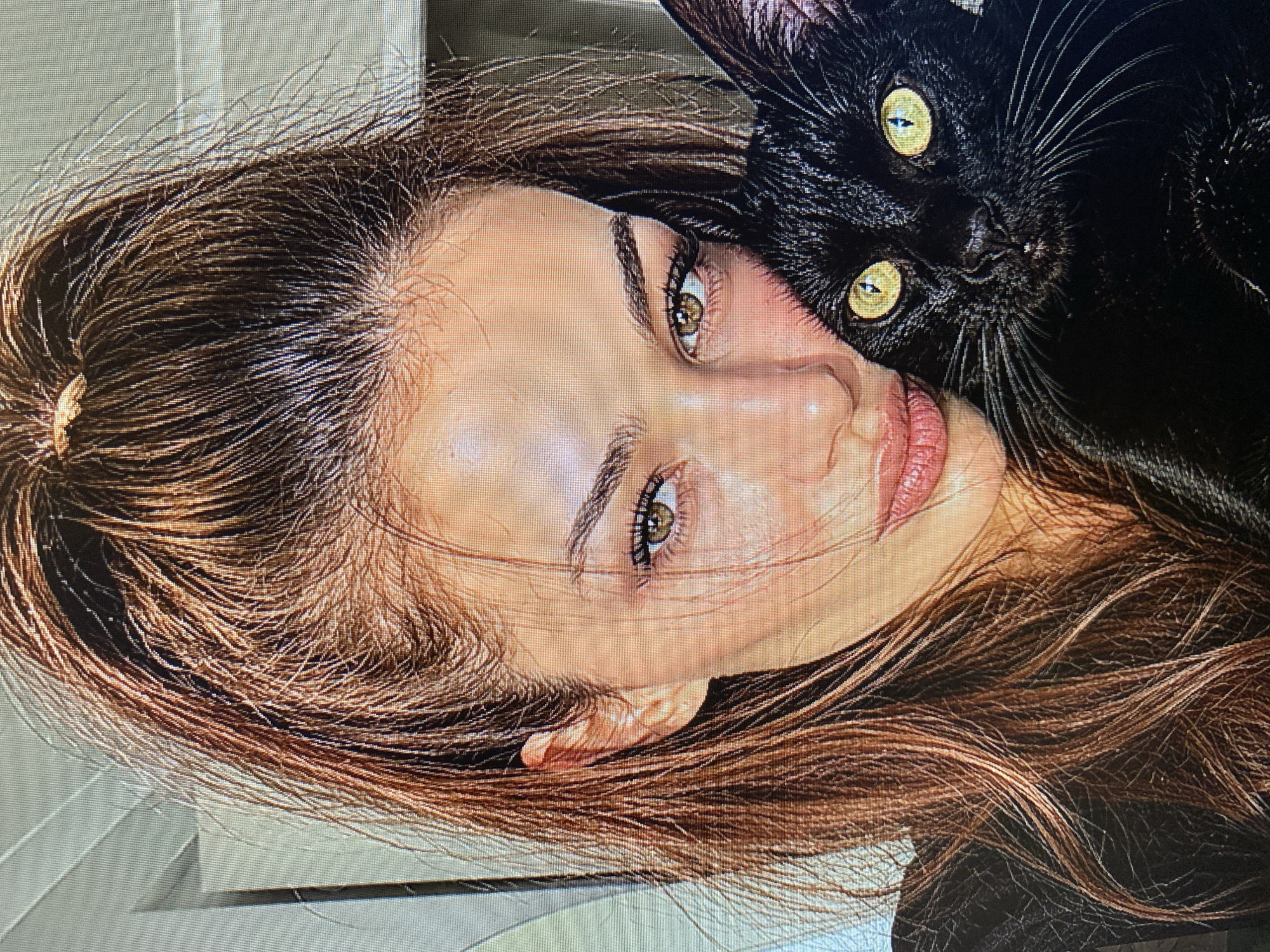 A close-up, waist-up portrait shows a young woman with long, auburn hair pulled into a high ponytail, holding a black cat. The woman is looking towards the camera with her head tilted slightly. She has striking green eyes and wears a subtle makeup. The cat, with piercing yellow eyes, is positioned in front of the woman's chest, with its head peeking over her shoulder and towards the viewer. The background is a plain, light-colored wall with some vertical lines. The image appears to be taken indoors. There is no discernible location in the city of Mahilyow, Belarus.