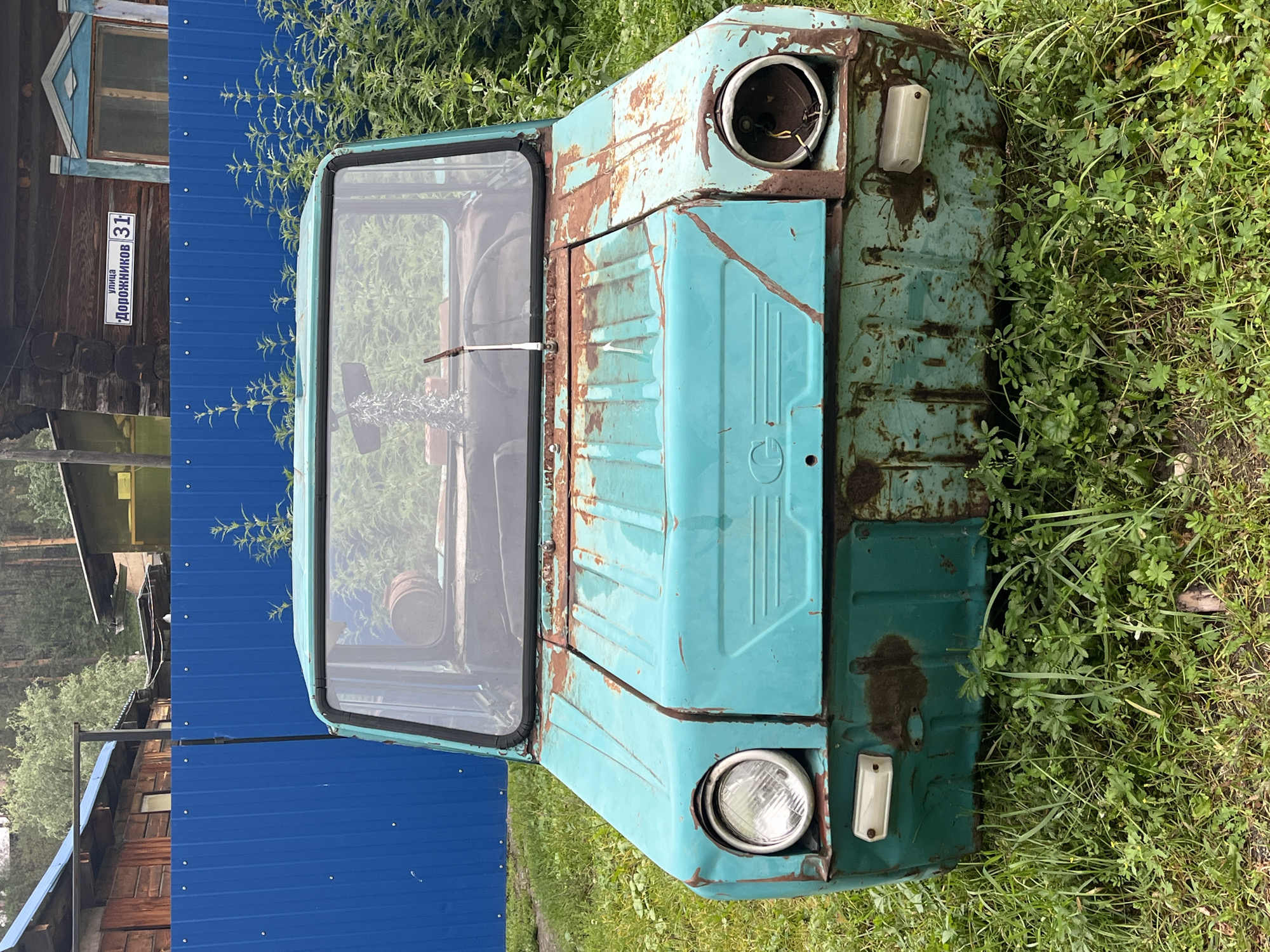The image captures a weathered, turquoise-colored vehicle, likely a microcar or utility vehicle, parked outdoors in what appears to be a rural or semi-rural setting. The car is heavily rusted, particularly on its hood and front fascia, with exposed metal showing orange-brown corrosion. Its headlights are intact, but one of the front grilles is damaged, and the bumper area is also showing significant rust. The vehicle's windows are visible, revealing the steering wheel and parts of the interior, which seems sparse. A decorative, possibly metallic or fabric, hanging ornament is visible from the rearview mirror. The car is positioned on a grassy area, with various types of green vegetation growing around and beneath it.
Behind the car, a bright blue corrugated metal fence runs horizontally across the frame. Beyond the fence, several structures are visible. To the left, a wooden building with a dark roof and exposed beams can be seen. To the right of this, another building with a similar dark roof and lighter-colored walls is present, partially obscured by foliage. Further in the background, more buildings with wooden construction and roofs are discernible, suggesting a residential or utilitarian area. A sign is affixed to one of the wooden buildings on the right, which, according to information provided about the location, reads "улица Дорожников 31" (Dorozhnikov Street 31), indicating the approximate location in Ust-Sema, Russia. The overall environment suggests an overcast or slightly diffused lighting condition, consistent with a temperate climate. No people are present in the image.