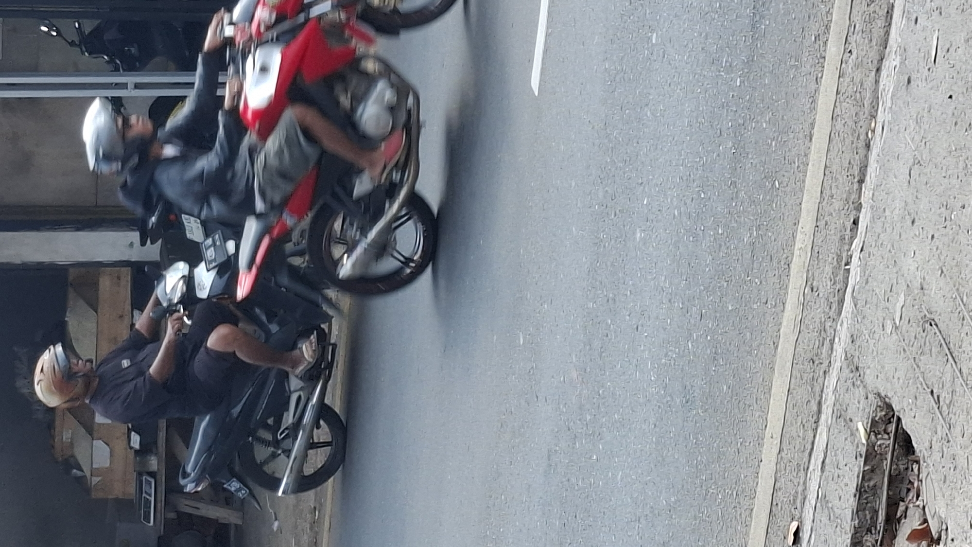 In the city of Cilaku, Indonesia, two individuals are captured on motorcycles in motion. The foreground shows a paved road with a distinct white line on the right side and a cracked concrete curb with exposed rebar on the left. Behind the curb, a section of the road appears to be under repair. In the middle ground, a man wearing a black t-shirt, black shorts, and a light-colored helmet is riding a black motorcycle. He is positioned on the left side of the image. To his right, a second individual, also on a motorcycle, is partially visible. This person wears a dark hoodie, light-colored shorts, and a silver helmet. The motorcycle they are riding is red. Both individuals are actively engaged in riding their motorcycles. The background depicts a building with vertical elements, possibly doors or windows, and a wooden structure behind the first motorcycle.