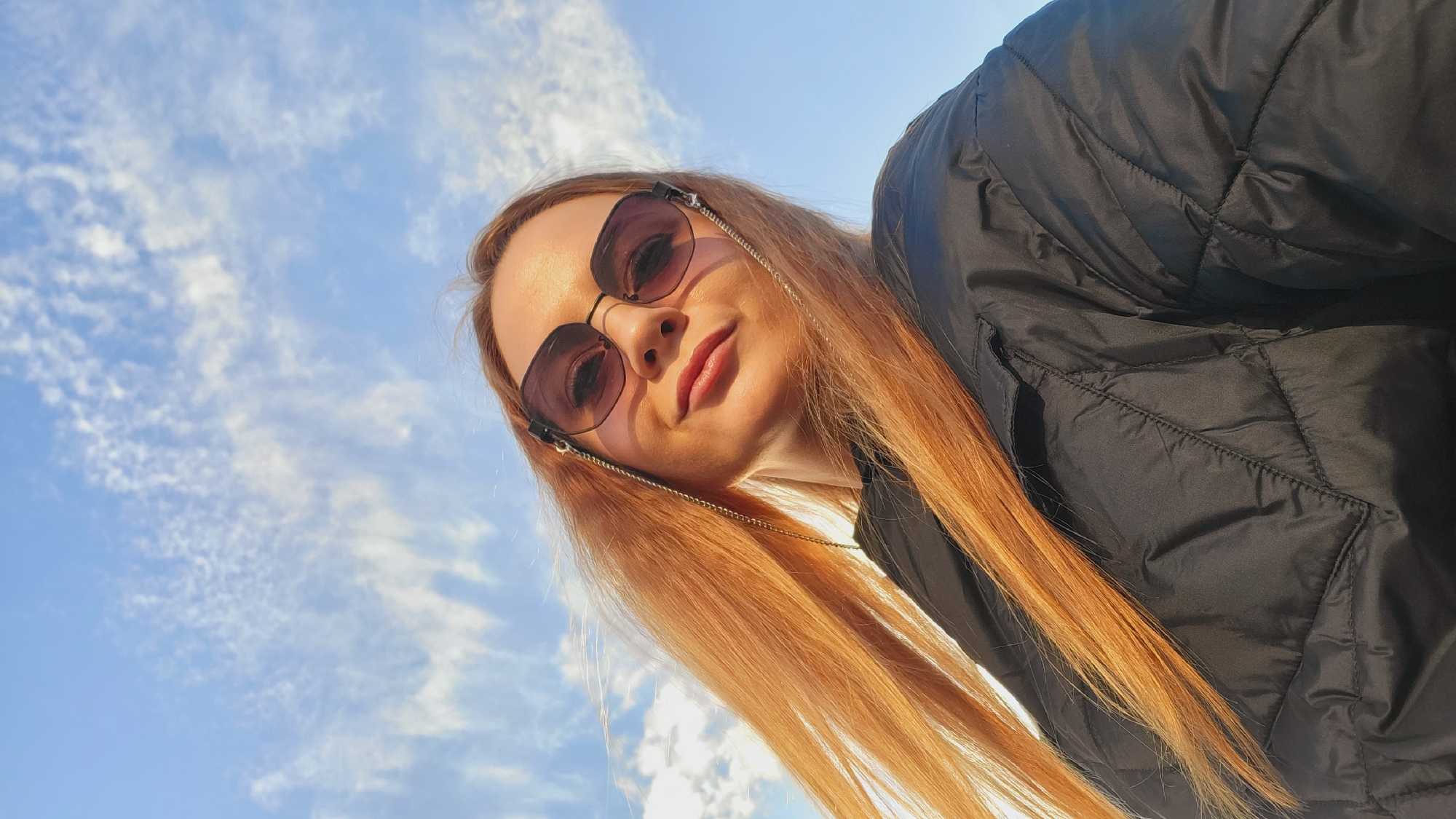 The image is a low-angle shot of a woman with long, straight, light-red hair, wearing sunglasses, and a black quilted jacket. The background is a blue sky with wispy clouds. It is not possible to determine the location within the city of Chaikovsky, Russia from the image. There is only one person in the image and no interactions visible.