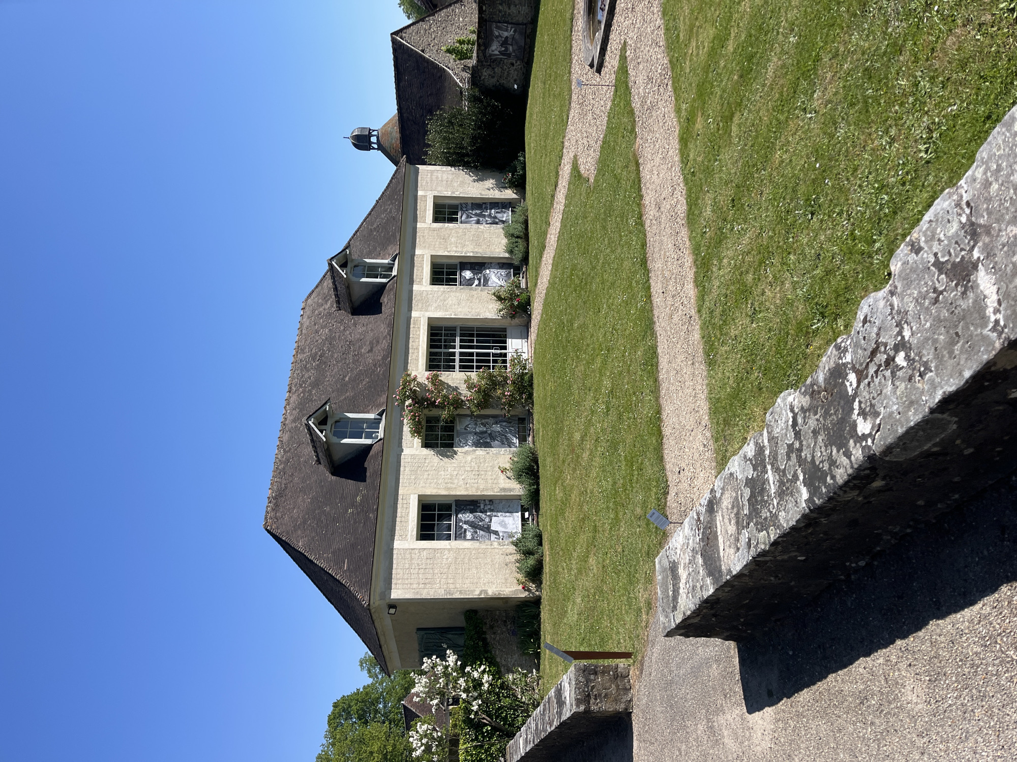 An outdoor, horizontal shot captures a historic-looking building set within a well-maintained garden under a clear blue sky. The building, constructed from light-colored stone or stucco, features a steep, dark-tiled roof with several prominent dormer windows. A small, dark cupola or bell tower rises from the roofline on the left side of the building.
The main facade of the building displays multiple windows, some of which appear to have black and white images or prints displayed behind the glass, possibly depicting historical figures or scenes. Climbing plants, including what looks like a rose bush, adorn the walls around some of the windows. Lush green grass covers the garden, traversed by a winding gravel path that meanders towards the background. In the immediate foreground on the right, a textured stone wall, covered with lichen, runs diagonally, casting a long shadow.
The scene is bathed in bright sunlight, suggesting midday or early afternoon, with distinct shadows visible on the grass and from the stone wall. The weather is clear and sunny, indicated by the vibrant blue sky. No people are present, lending a tranquil atmosphere to the setting. The architectural style and garden design are characteristic of a European estate or manor, consistent with the location of Saint-Rémy-lès-Chevreuse, France. Small, unreadable white plaques are visible on the grass and near the stone wall.