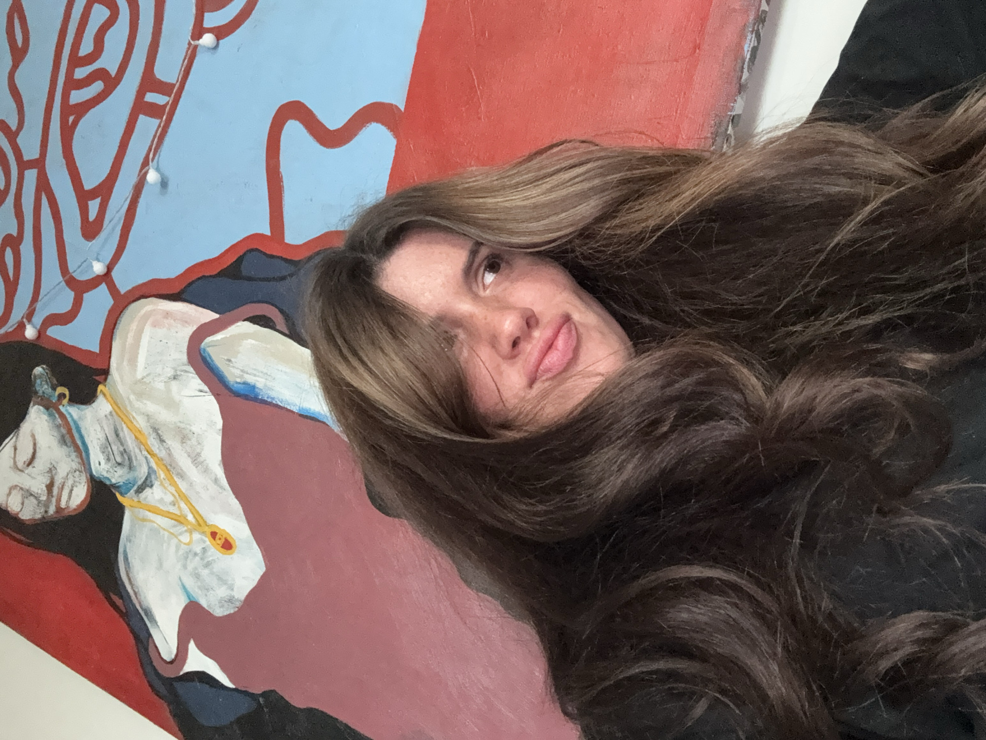 A close-up, tilted portrait captures a woman with long, wavy dark brown hair featuring lighter highlights. Her face, partially visible from the forehead to the chin, displays a playful expression with slightly pursed lips and visible freckles. The woman is positioned against a vibrant, abstract painting or mural.
The artwork behind her consists of several distinct sections. In the upper left, abstract red lines are drawn over a light blue background. To the right of this, a large section of the painting is a solid, bright red. Below these, a stylized figure or object is depicted in shades of white, black, and maroon/dark red. This figure appears to be adorned with a golden necklace or chain, which has a small red pendant. A string of small white fairy lights drapes across the light blue and red abstract sections of the painting.
The setting is indoors, likely a casual space such as a home or artist's studio, providing a colorful backdrop for the portrait. The image appears to be a selfie or a close-up photograph. No specific time of day, weather conditions, or visible text can be determined from the image. The general location context is Krasnoyarsk, Russia.