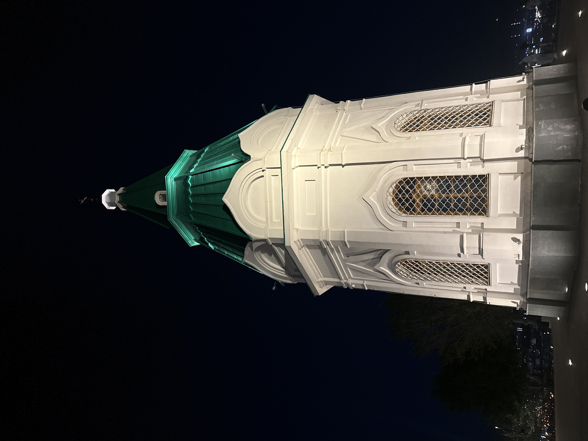 The image shows the illuminated dome of a white building against a dark night sky. The dome is green and has a pointed, architectural design. The white part of the building features ornate detailing, including arched windows with intricate lattice work. Some lights are visible on the white surface, and there are also some lights on the darker background, suggesting a cityscape. The time of day is clearly night due to the darkness of the sky and the artificial lighting. There is no visible text in the image. The scene is static, with no obvious activity taking place. The location is specified as Krasnoyarsk, Russia.