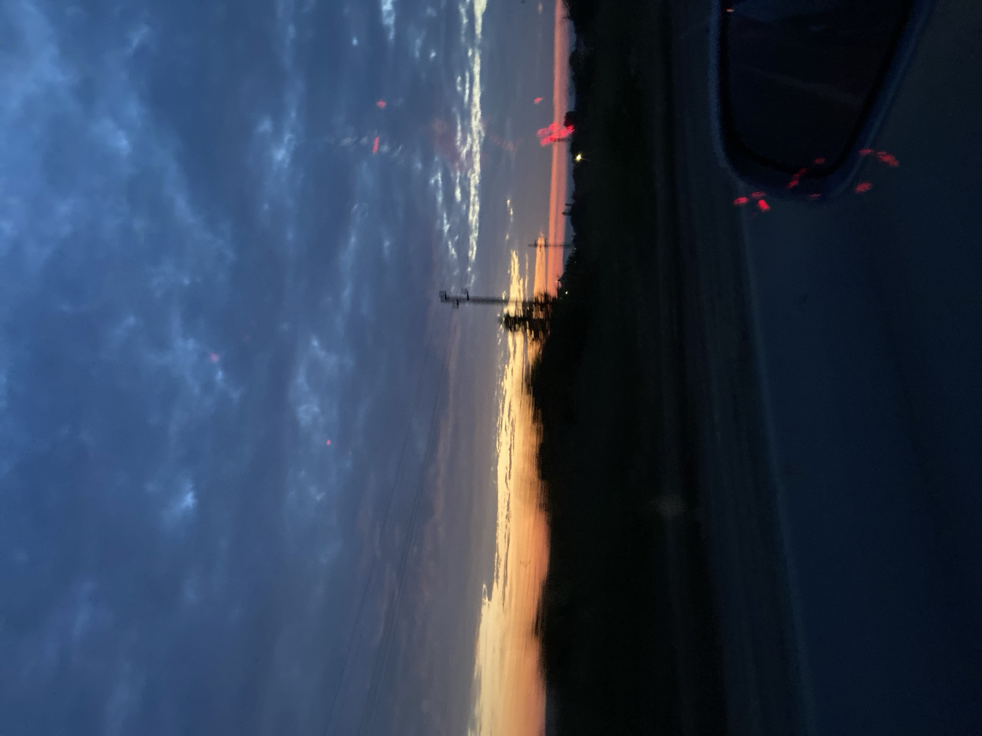The image displays a twilight sky, likely at sunset, captured from within a moving vehicle. The sky dominates the upper portion of the frame, featuring dark blue and grey clouds, some of which are illuminated by the setting sun. A vibrant horizontal band of orange and yellow light stretches across the horizon, casting a warm glow and highlighting the undersides of the clouds.
Below this colorful horizon, the landscape is silhouetted in deep, dark tones, appearing as a strip of trees or dense foliage. A utility pole, possibly for power or telephone lines, stands on the left against the glowing sky, with faint wires extending across the scene.
On the far right, a dark, blurred shape indicates the interior of the vehicle, possibly a window frame or dashboard. Several small, bright red, blurred lights are scattered across the sky area and clustered within the vehicle's interior, likely reflections or distant vehicle taillights. The overall blur within the image suggests it was captured while the vehicle was in motion. There is no visible text. The scene depicts a drive through a rural or semi-rural area during dusk.