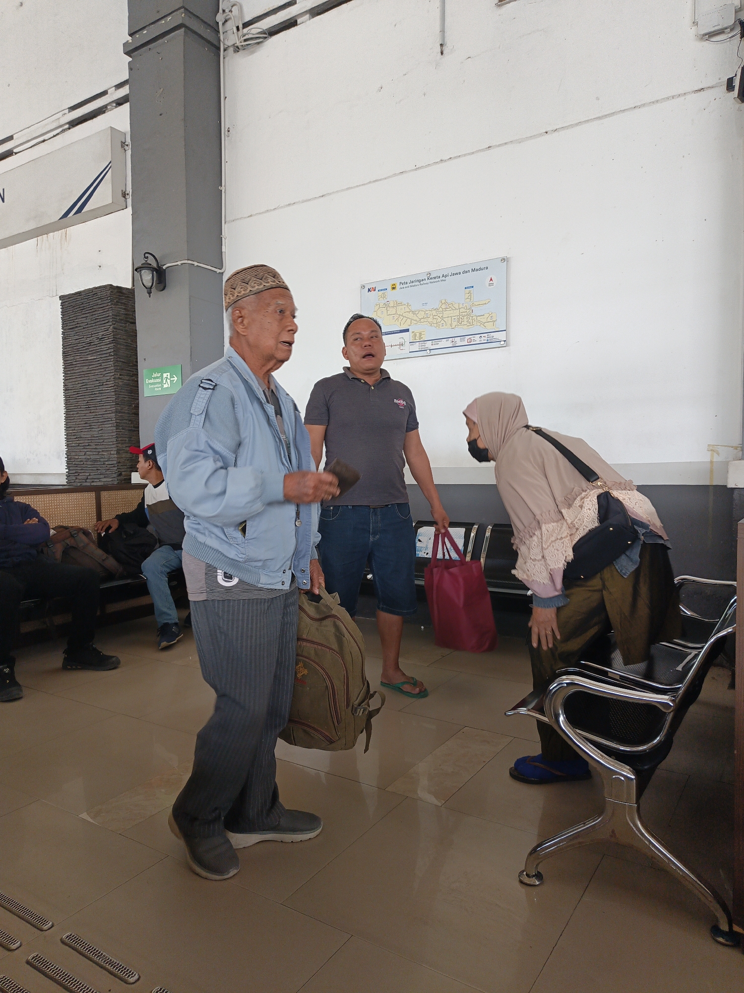 The image depicts an indoor scene, likely a waiting area in Klaten, Indonesia. Three people are the central focus. An elderly man in a light blue jacket and a patterned cap stands facing to the right, holding a dark object in his right hand and a large backpack over his left shoulder. Beside him and slightly behind, a man in a gray t-shirt and blue shorts is looking towards the right with his mouth open. To the right of this man, a woman wearing a beige hijab and a long-sleeved top is bending down. She is wearing a black face mask. In the background, a map titled "Peta Jaringan Kereta Api Jawa dan Madura" (Java and Madura Railway Network Map) is displayed on a white wall. Several empty chairs with metallic frames are visible. Other individuals are seated in the background on benches. The floor is tiled.