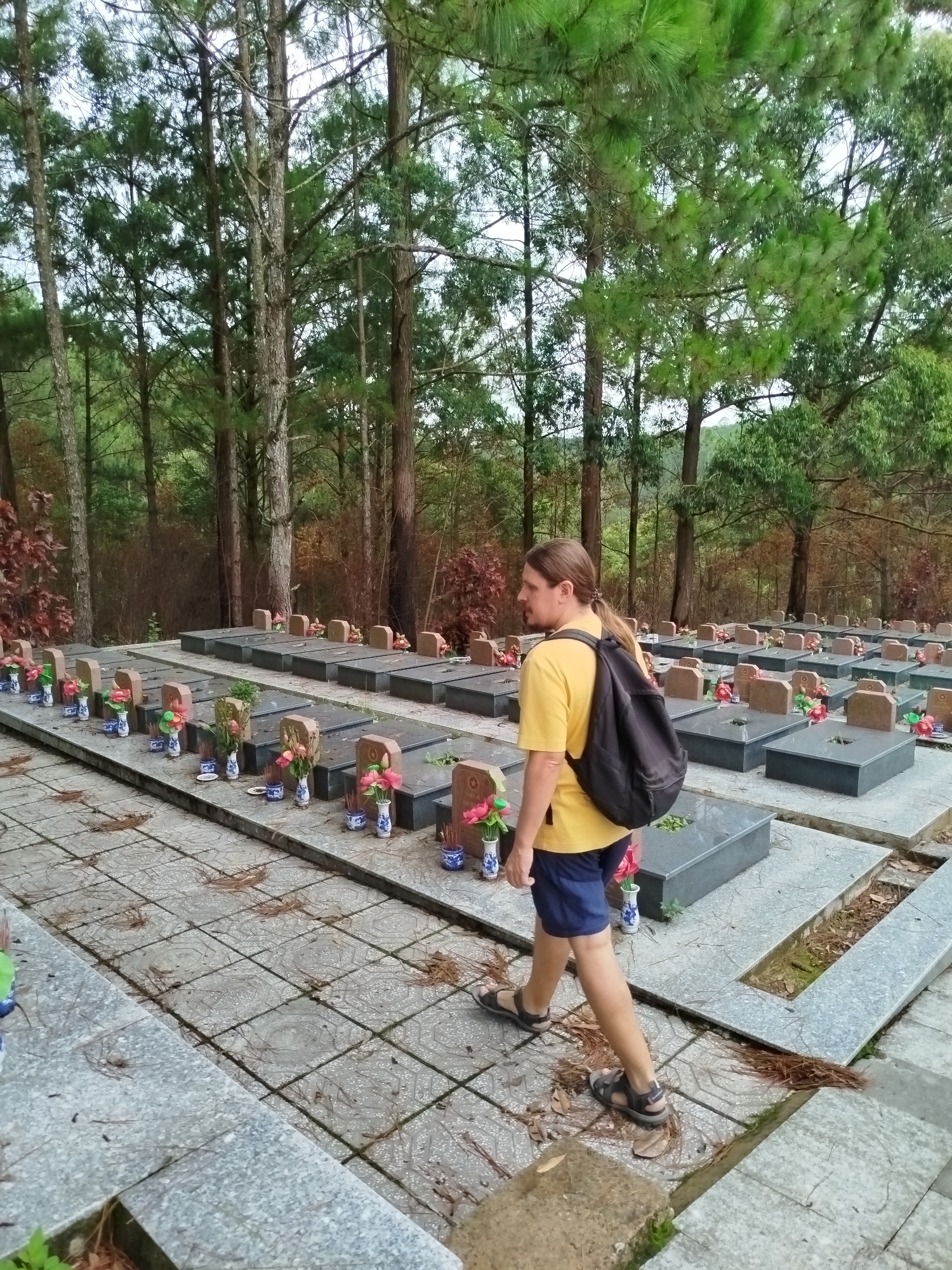 A man with long brown hair tied back, wearing a yellow t-shirt, dark shorts, and sandals, walks through a cemetery. He carries a black backpack and is seen in profile, moving from left to right across a tiled path. The path is covered with pine needles and leaves.
The cemetery features numerous rectangular graves or tombs arranged in rows on elevated platforms. These graves are made of grey and dark grey stone, with some featuring small, light brown markers. Many graves are adorned with small, ornate blue and white vases containing artificial pink and orange flowers, and some also have incense sticks or offerings.
The cemetery is set within a dense forest of tall, slender pine trees, creating a shaded and natural environment. In the background, some trees display reddish-brown foliage. The overall lighting suggests it is daytime, possibly overcast, contributing to a subdued atmosphere. The location is consistent with Cam Ly - Đà Lạt, Vietnam. There is no visible text in the image.