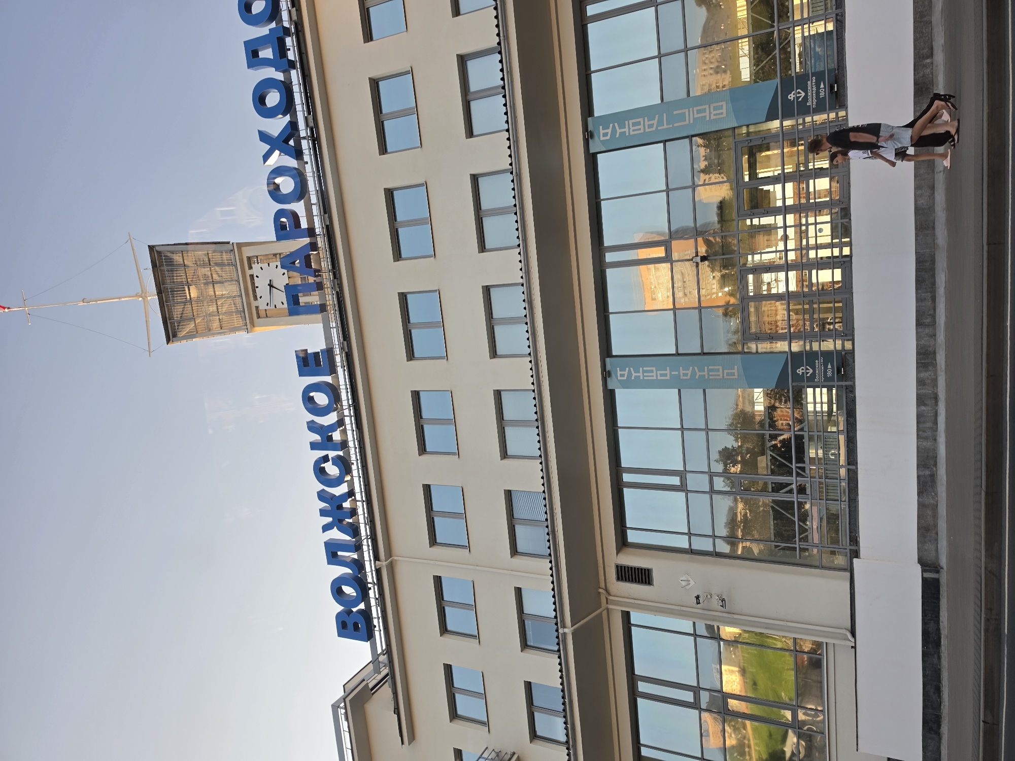 The image shows the building of the Volga River Shipping Company in Nizhny Novgorod, Russia. The building is a modern, multi-story structure with large glass windows. A large sign with blue lettering on the top of the building reads "ВОЛЖСКОЕ ПАРОХОДСТВО," which translates to "VOLGA SHIPPING COMPANY." A clock tower with a flag is visible on the right side of the building. In front of the building, two people are walking on the sidewalk. One person appears to be a woman wearing a black shirt and shorts, and the other is a younger person wearing a white shirt and dark shorts. The reflection of buildings and trees can be seen in the glass windows. The sky is clear and blue.