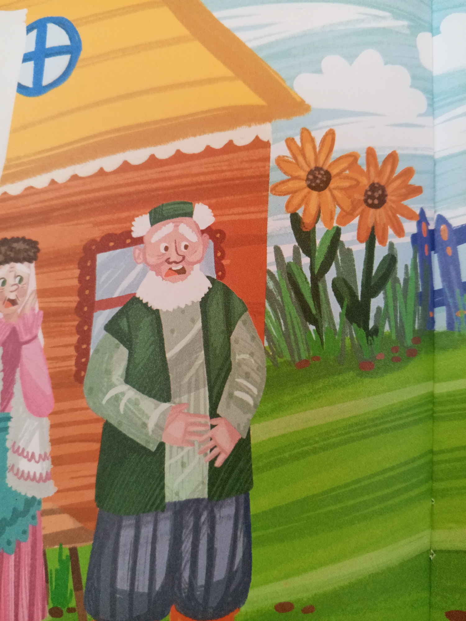 The image depicts a stylized illustration from a children's book, featuring a whimsical scene with two elderly characters standing outside a quaint, cartoonish house. The house has an orange-brown wooden exterior and a yellow roof adorned with a blue circular window that has a cross-like shape within it. To the left of the house, a woman with dark curly hair and a surprised expression wears a pink dress and a white ruffled collar. To her right, an older man with white hair and a beard, wearing a green cap, a green tunic over a lighter grey garment, and dark striped pants, gestures with his hands. In the background, two large orange flowers bloom amidst tall green stalks, and a blue picket fence encloses a portion of the scene. The ground is a vibrant green, with streaks indicating rolling hills. The sky above is a light blue with white, fluffy clouds. The image does not provide any specific visual information that would allow for its localization to the city of Chistopol, Russia.