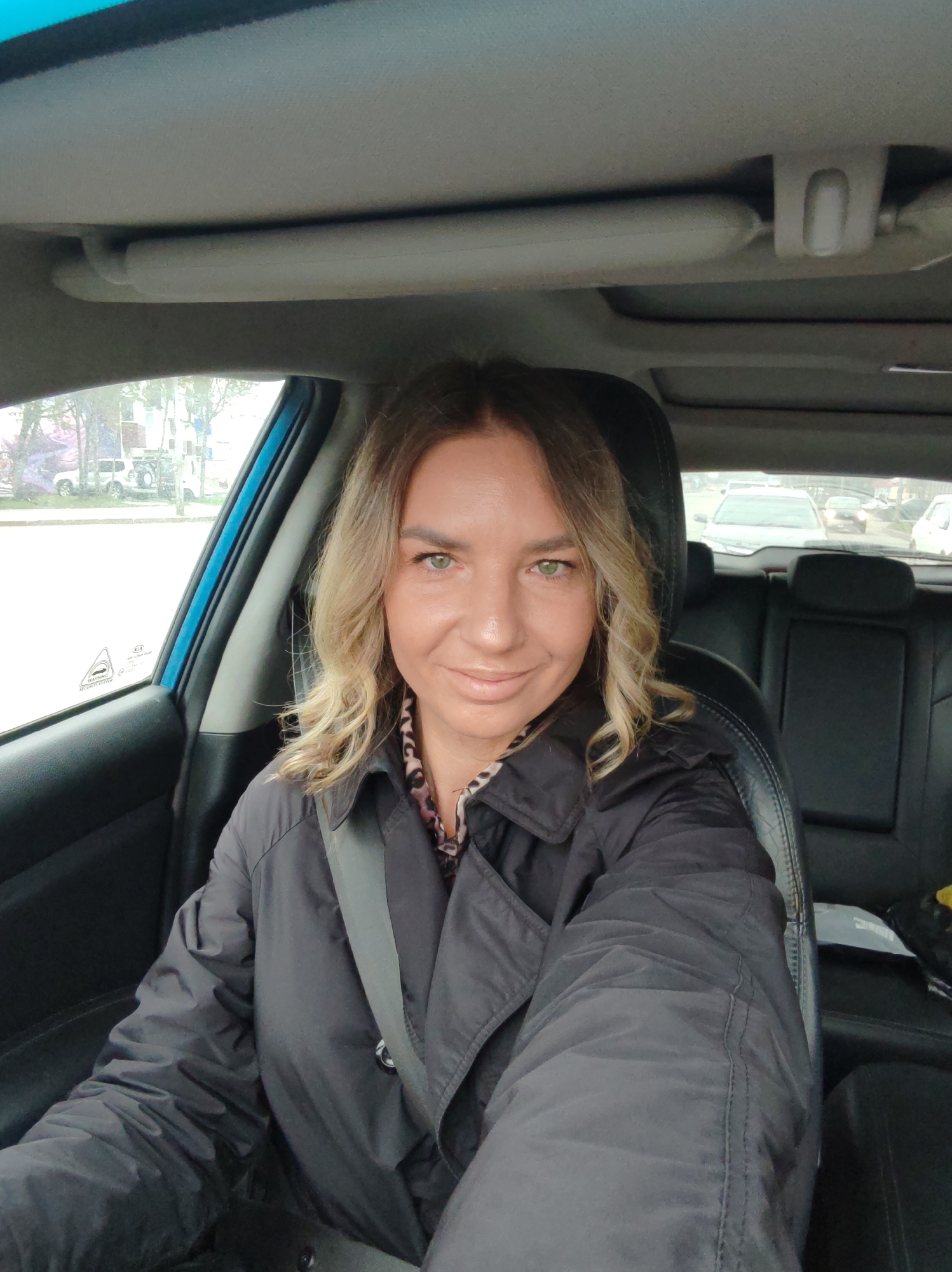 A woman is seen in a selfie, sitting in the driver's seat of a car, likely in Petropavlovsk-Kamchatsky, Russia. She is smiling faintly at the camera, with shoulder-length wavy blonde hair and light green eyes. She is wearing a black collared jacket over a top with a subtle animal print pattern, and her seatbelt is fastened across her chest.
The car's interior features black leather seats and a dark dashboard. The light gray headliner, including a sun visor and an overhead console, is visible above her. Through the driver's side window, a street scene is blurred in the background, showing trees, a building with what appears to be a large poster or mural, and parked vehicles, including a white SUV. Visible text on the side window includes "KIA" and a "WARNING" label. Through the rear window, other cars can be seen on a road. The lighting indicates it is daytime, possibly with overcast weather conditions.