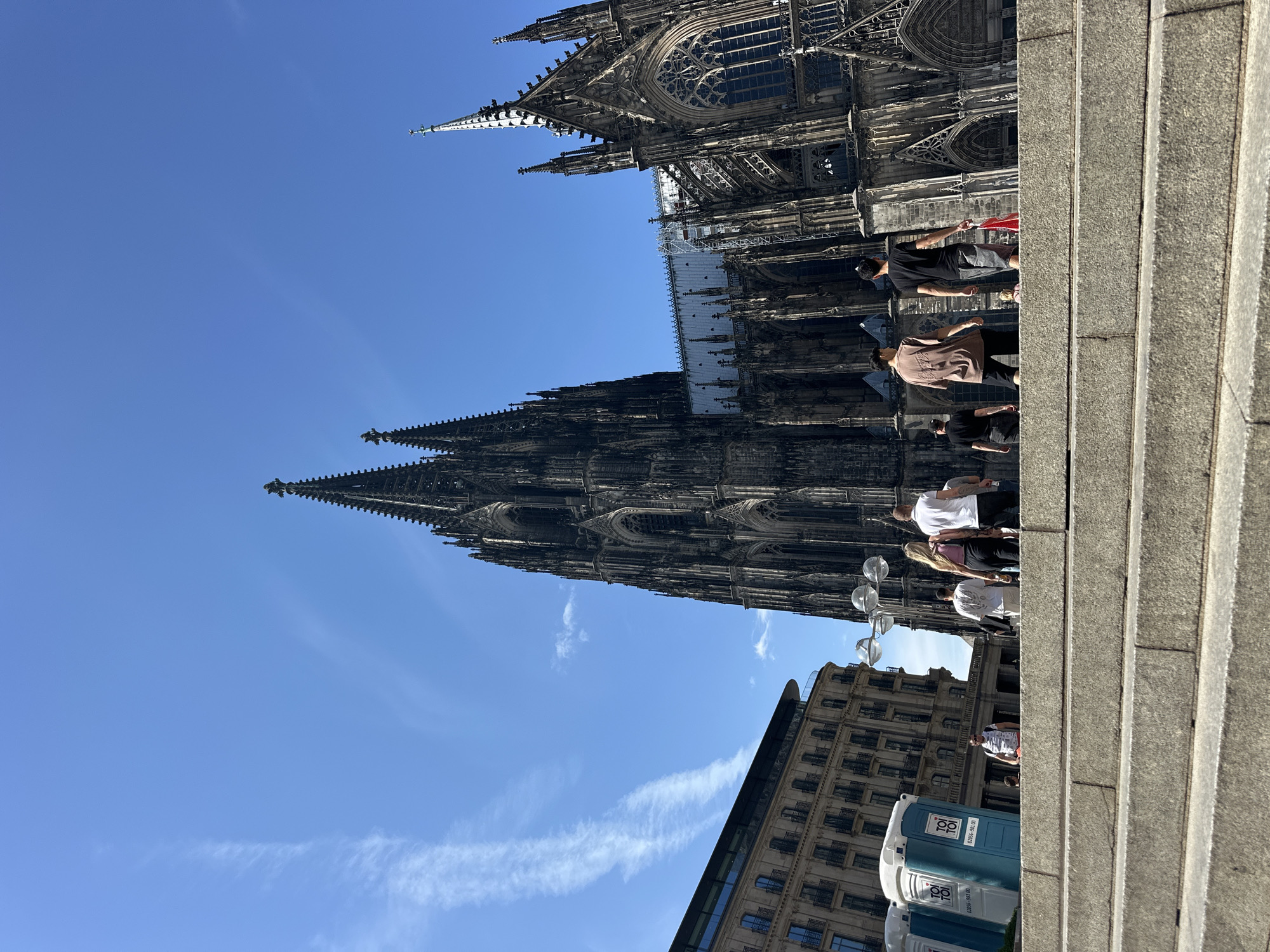 The image captures a daytime scene in Cologne, Germany, focusing on the Cologne Cathedral and its surroundings. The foreground features a wide, shallow stone staircase, with several individuals walking up and down. The people are casually dressed, with varied attire, and are seen from the back or side. To the left of the stairs are at least two blue portable toilets. In the mid-ground, the imposing Cologne Cathedral dominates the scene with its intricate Gothic architecture, pointed spires, and stained glass windows. Adjacent to the cathedral, on the left side of the image, is a multi-story building with a modern facade featuring numerous windows and balconies. The sky is a clear, bright blue with some wispy white clouds. The lighting suggests it is a sunny day.