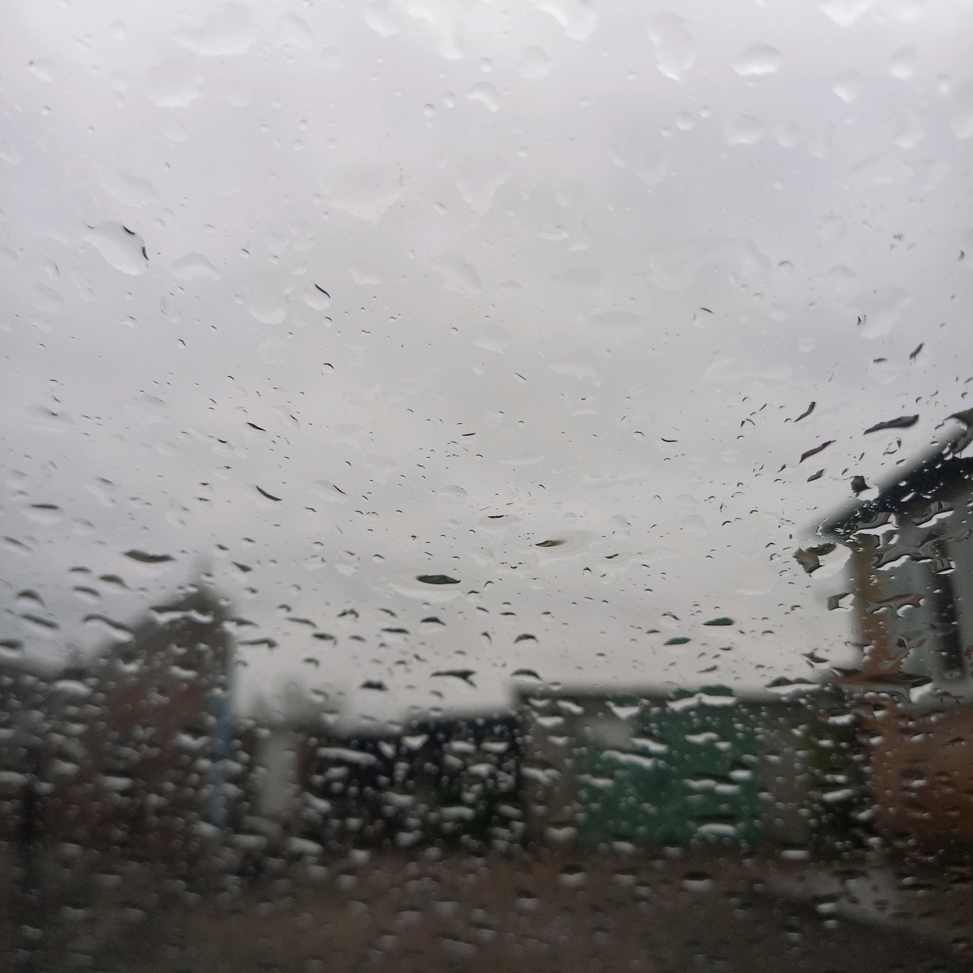 The image depicts a view through a window covered in raindrops, with the sky appearing overcast and grey. In the background, blurred structures that appear to be houses or buildings are visible, suggesting an outdoor scene in a residential area. One building on the right side of the image has a dark roof and light-colored walls. The overall impression is one of a rainy day. There is no indication of the city of Klyntsi, Ukraine within the image.